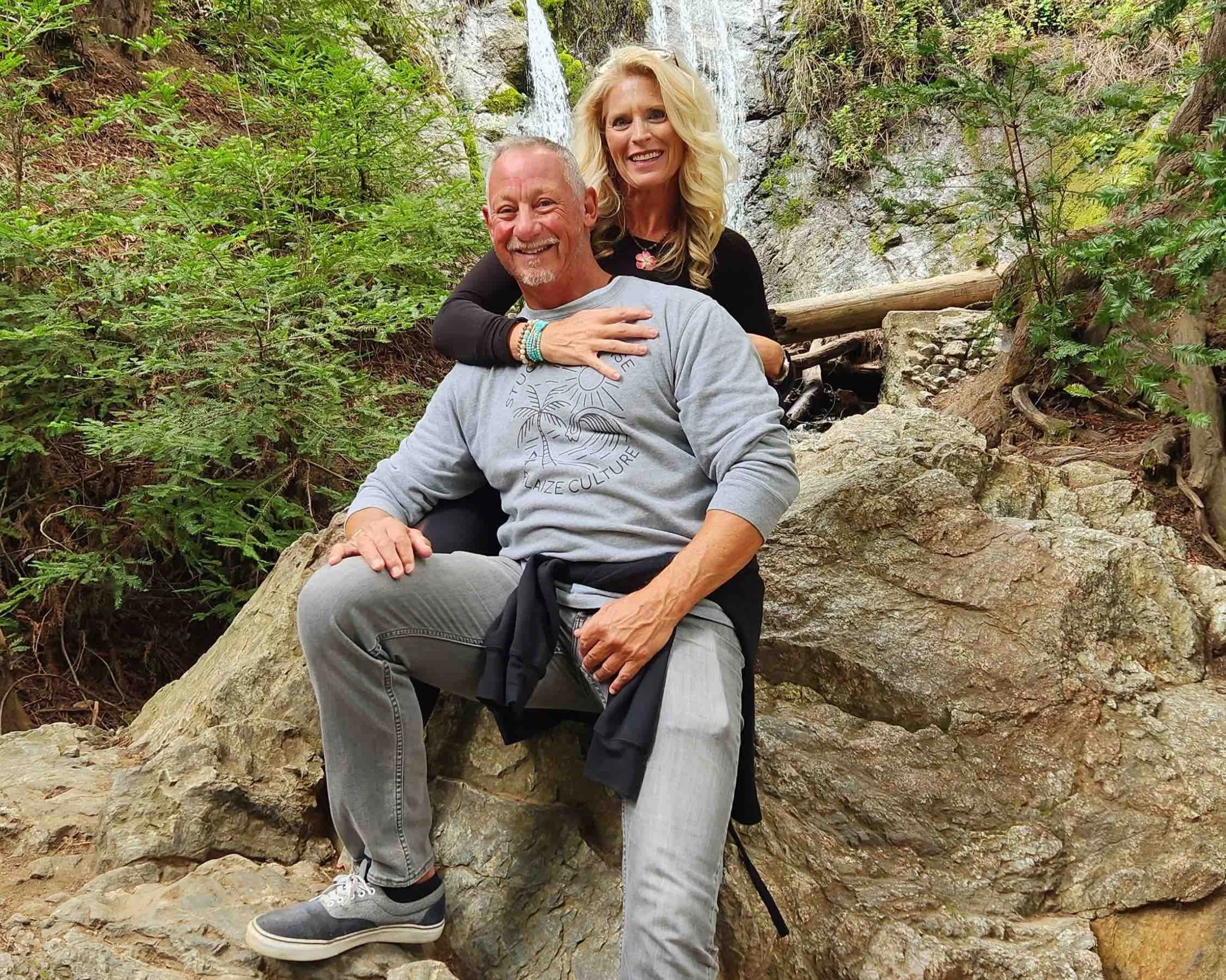 a woman embracing a seated man from behind on rocks with lush green trees around them