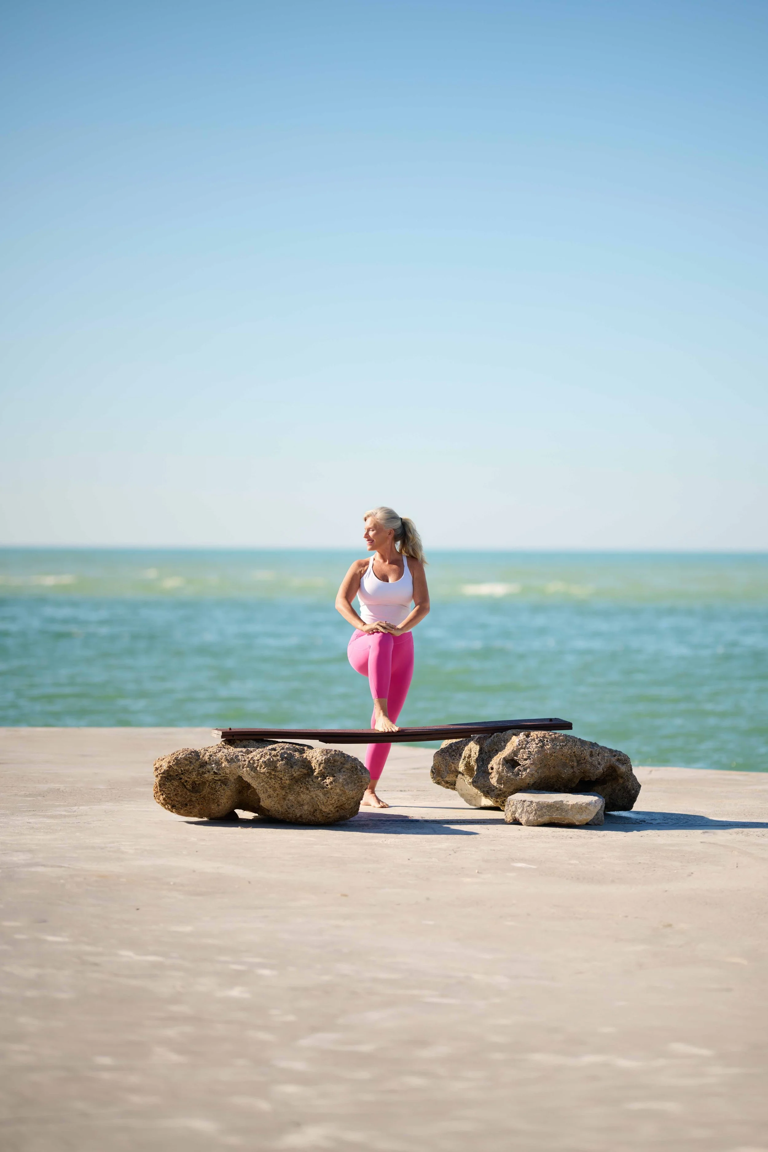 A woman practicing yoga on a beach, balancing on one foot on a balance beam situated on rocks, with the ocean and clear blue sky in the background.