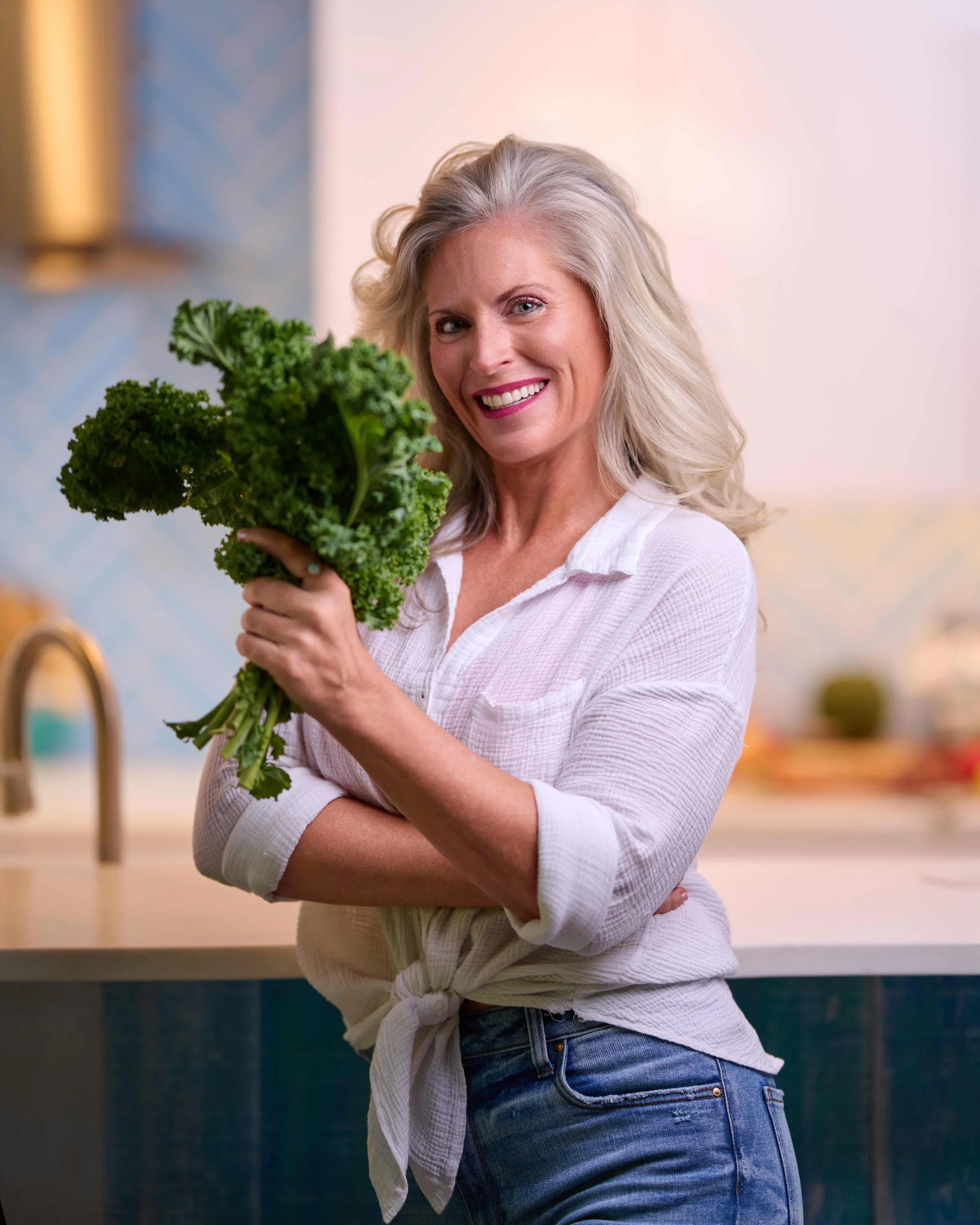 woman smiling at the camera in a white top and denim jeans while holding green kale