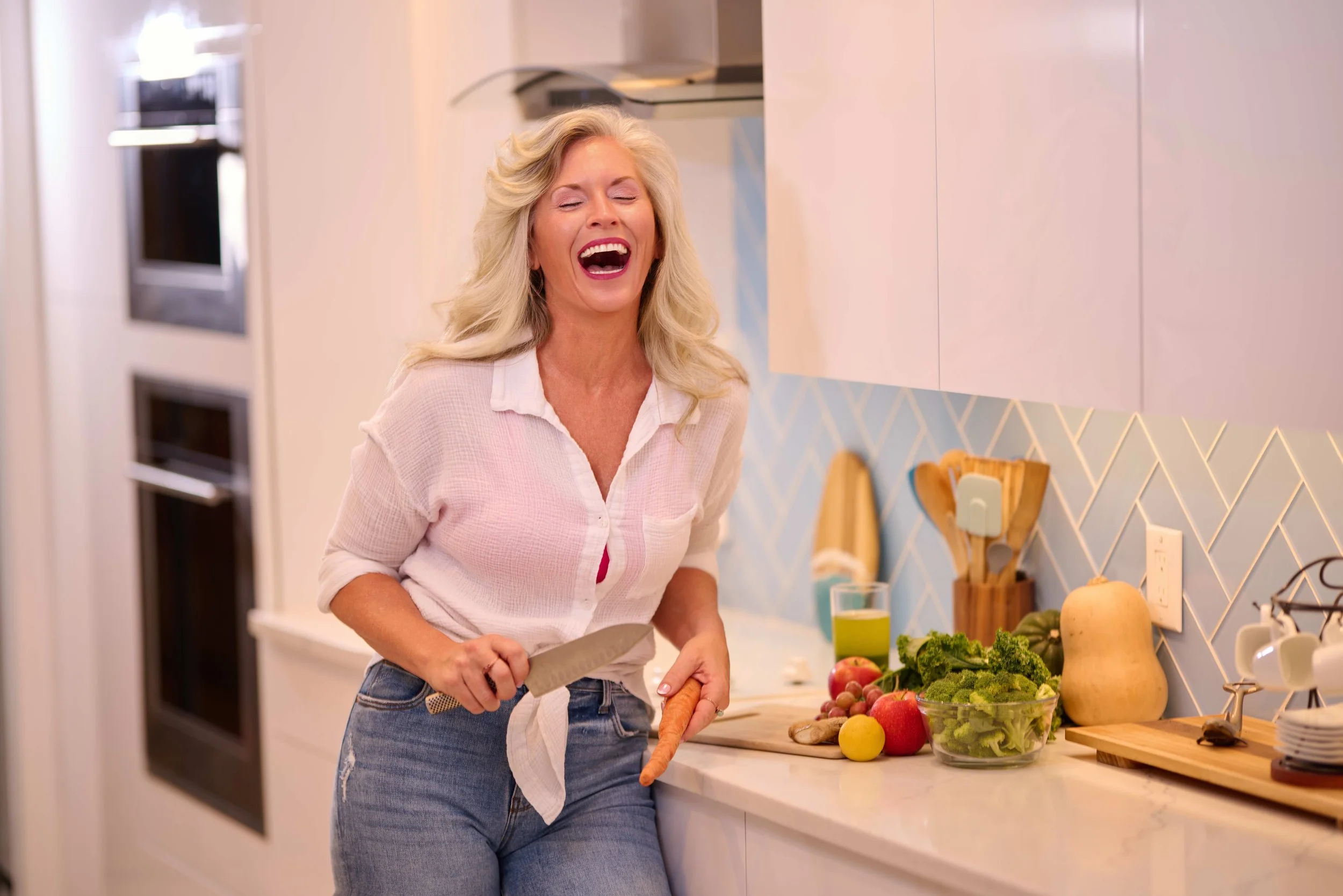 Woman with blonde hair laughing while preparing vegetables in a modern kitchen.