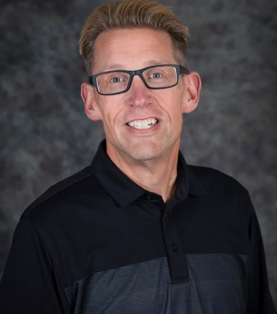 A middle-aged man with light brown hair, black glasses, wearing a black and dark gray polo shirt, smiling at the camera against a dark textured background.