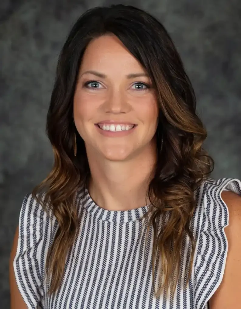 A woman with shoulder-length brown hair with subtle highlights, smiling, wearing a sleeveless black-and-white striped top with ruffled shoulders, against a dark gray textured background.