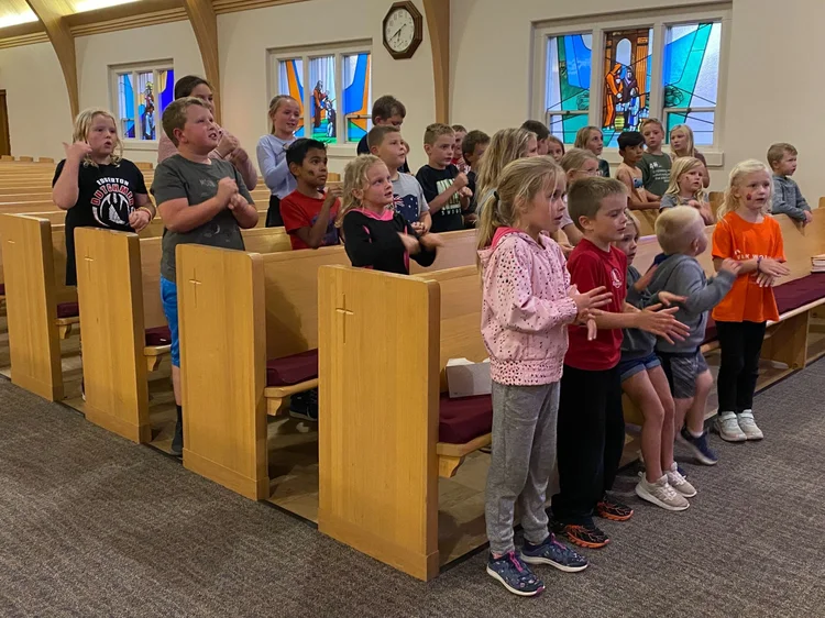 Group of children standing and clapping in church pews, stained glass windows in background.