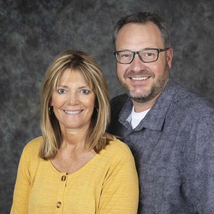 A woman with blonde hair wearing a yellow top and a man with glasses and gray hair wearing a gray shirt and blazer pose together smiling in front of a dark textured background.