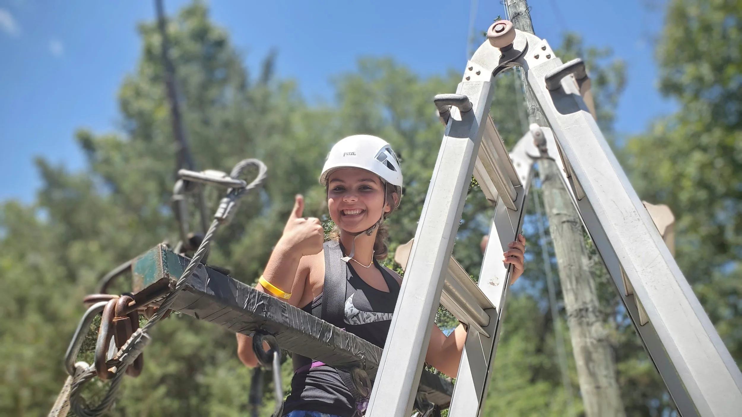 Person wearing a safety helmet and harness on a ladder, giving a thumbs-up with a blurred outdoor background.