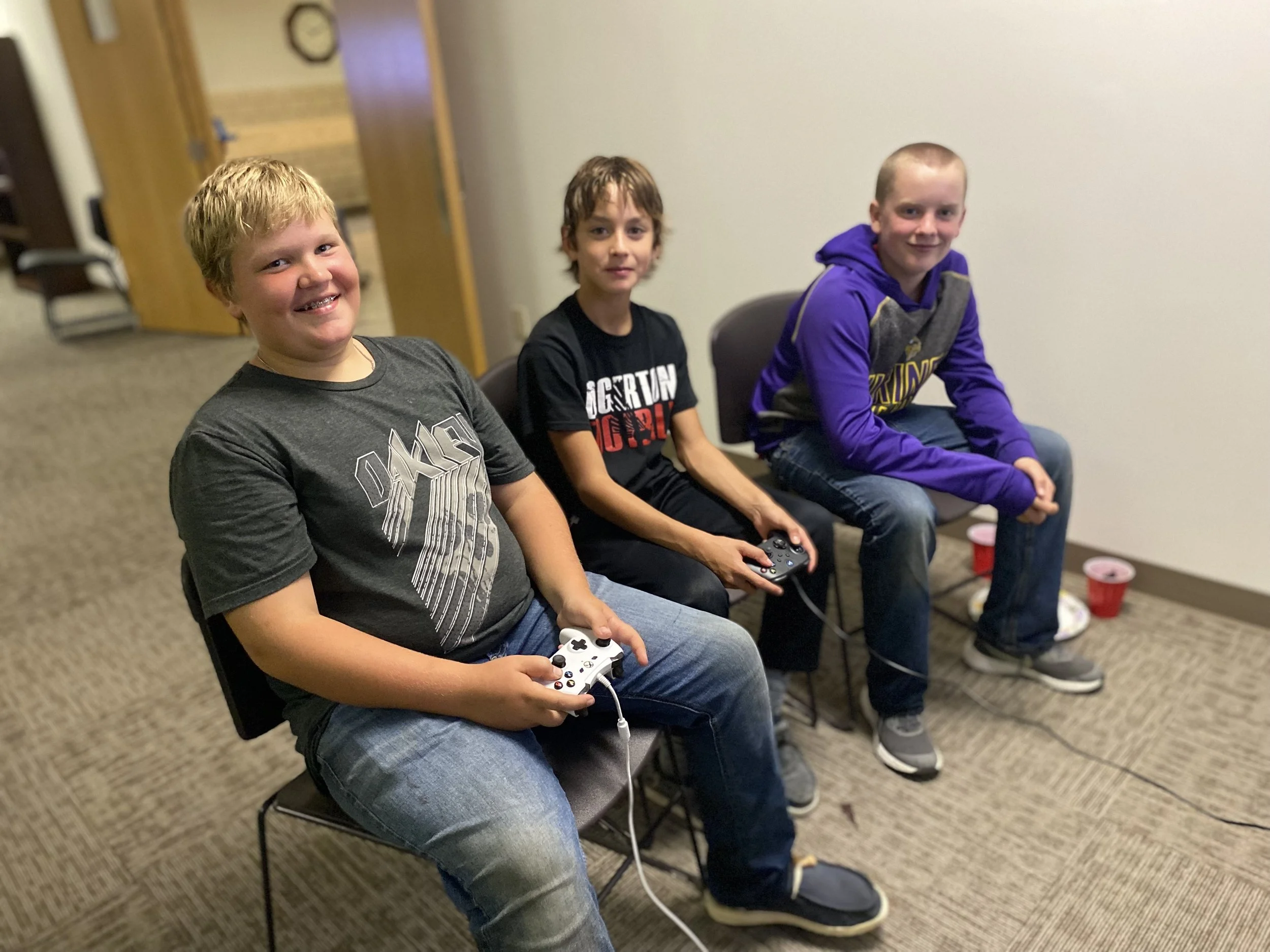 Three boys sitting on chairs in a room, holding video game controllers.