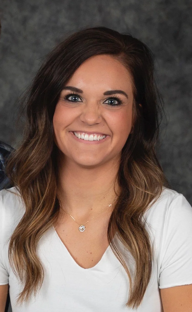 Smiling woman with brown hair, wearing a white shirt and a necklace, posing in front of a neutral background.