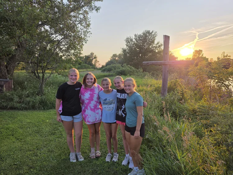 Students standing together in front of a cross at sunset