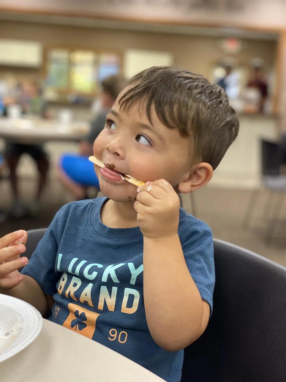 Young child licking a spoon in a casual dining setting, wearing a blue "Lucky Brand" T-shirt, with plates on the table.