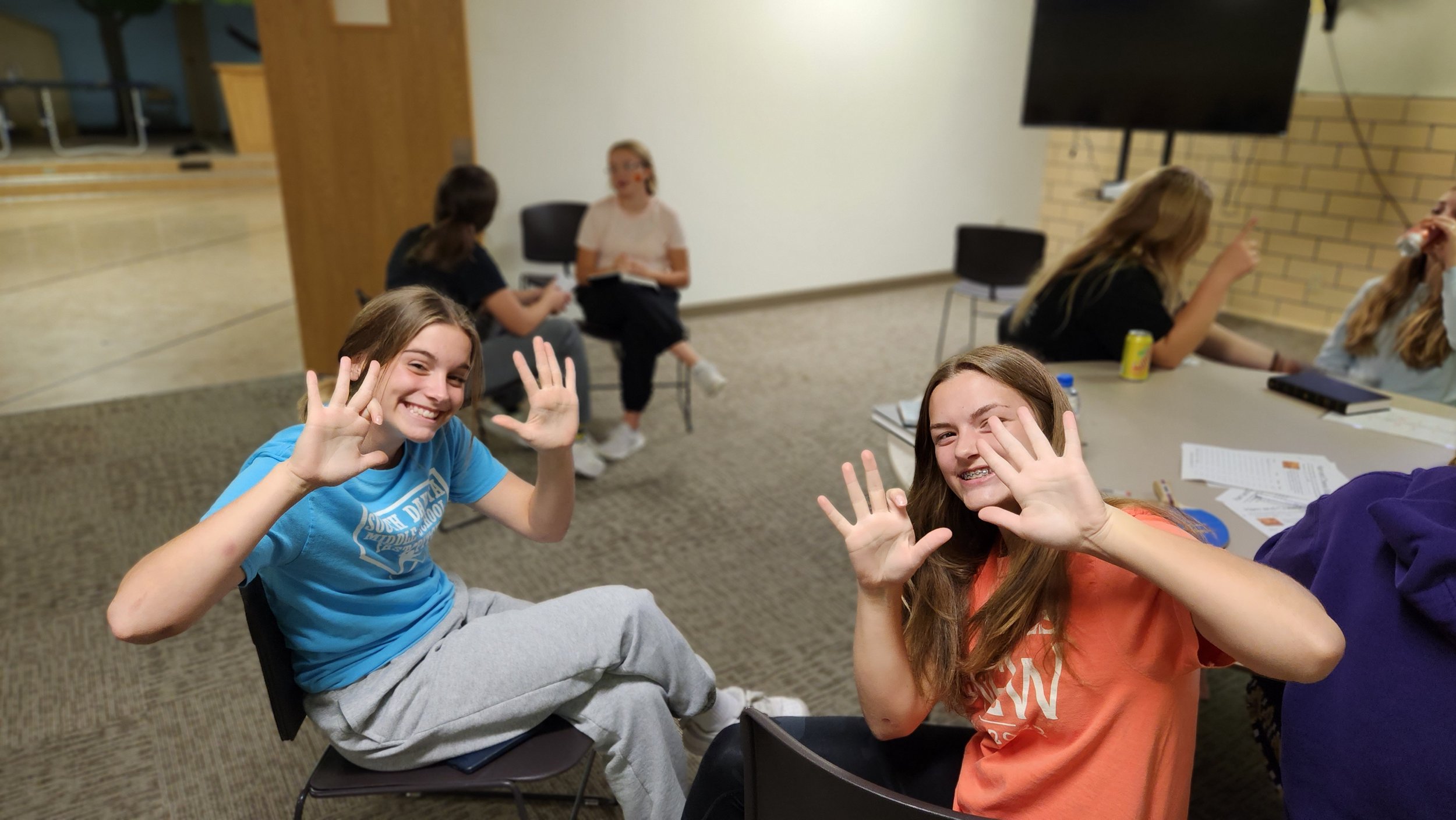 Group of young people sitting indoors, some smiling and gesturing at the camera. Two individuals in the foreground are prominently raising their hands. The setting appears to be a casual meeting or study session with papers and drinks on the table.