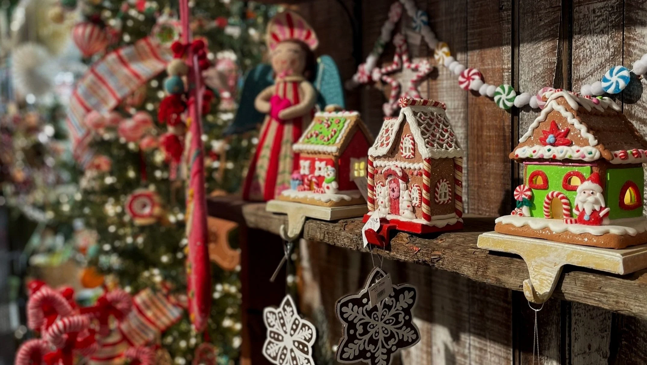 Display of decorated holiday gingerbread houses on a rustic wooden shelf with Christmas ornaments and tree in the background.