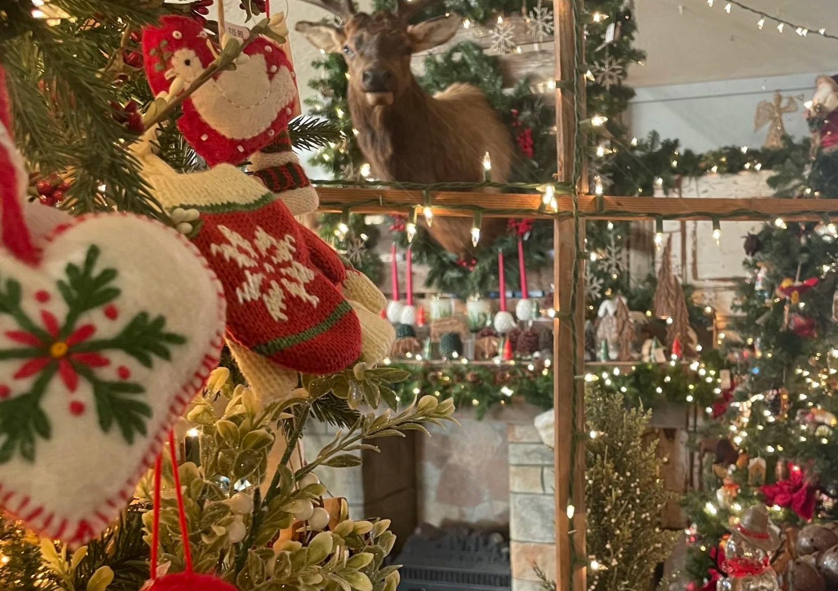 Close-up of Christmas stockings hanging on a decorated Christmas tree with an elf stocking in the foreground. In the background, a lit reindeer ornament is visible through a wooden shelf adorned with lights, showcasing a cozy holiday scene.