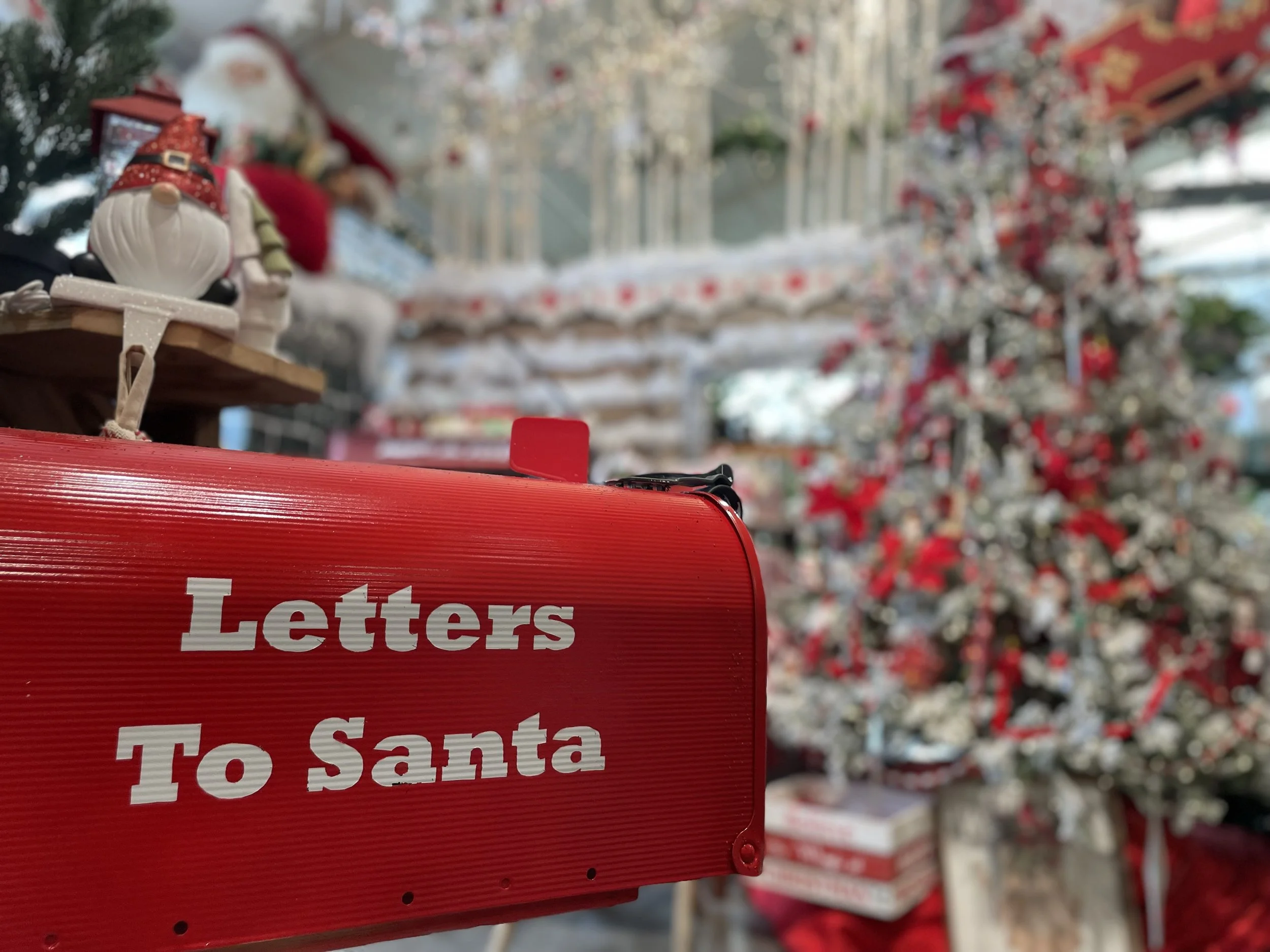 Close-up of a red mailbox labeled "Letters To Santa," with a decorated Christmas tree and holiday decorations blurred in the background.