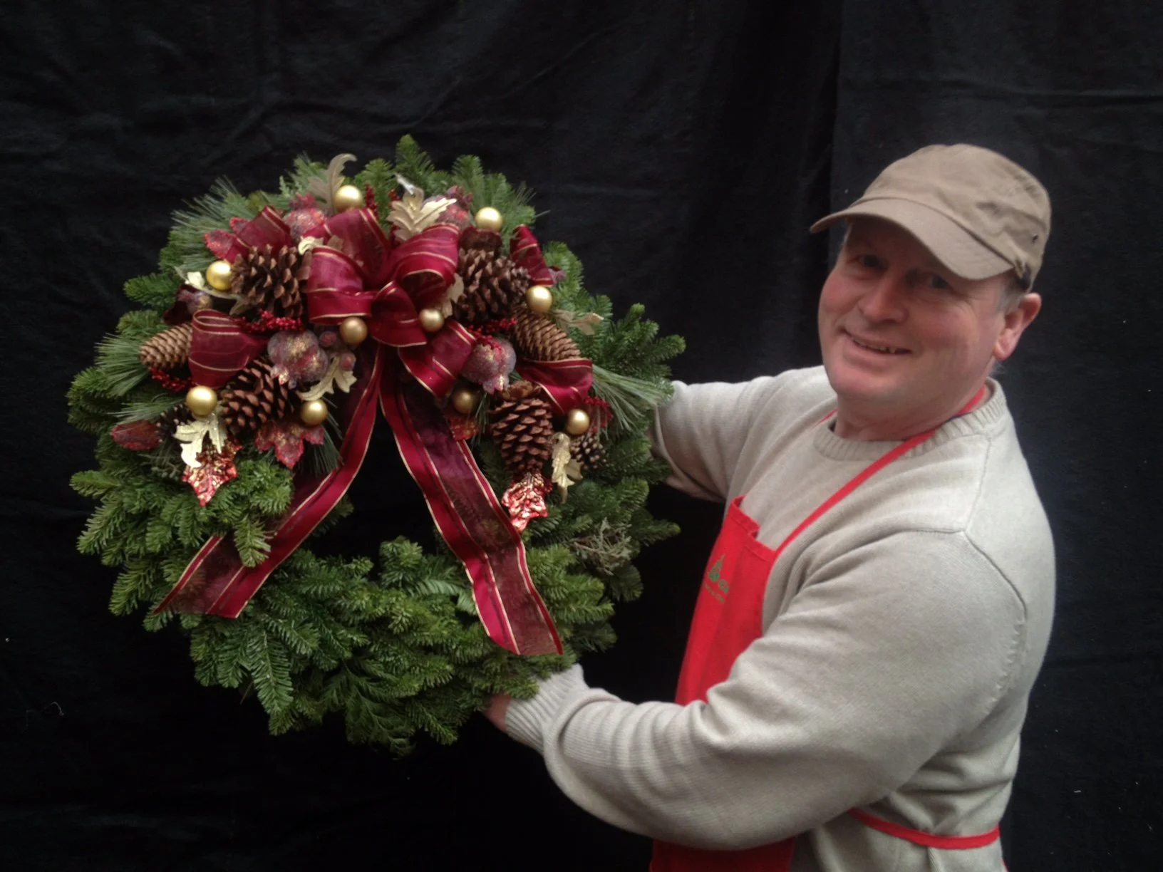 A man wearing a beige cap, light gray sweater, and red apron holding a Christmas wreath decorated with pine cones, gold ornaments, red ribbons, and holly leaves against a black background.