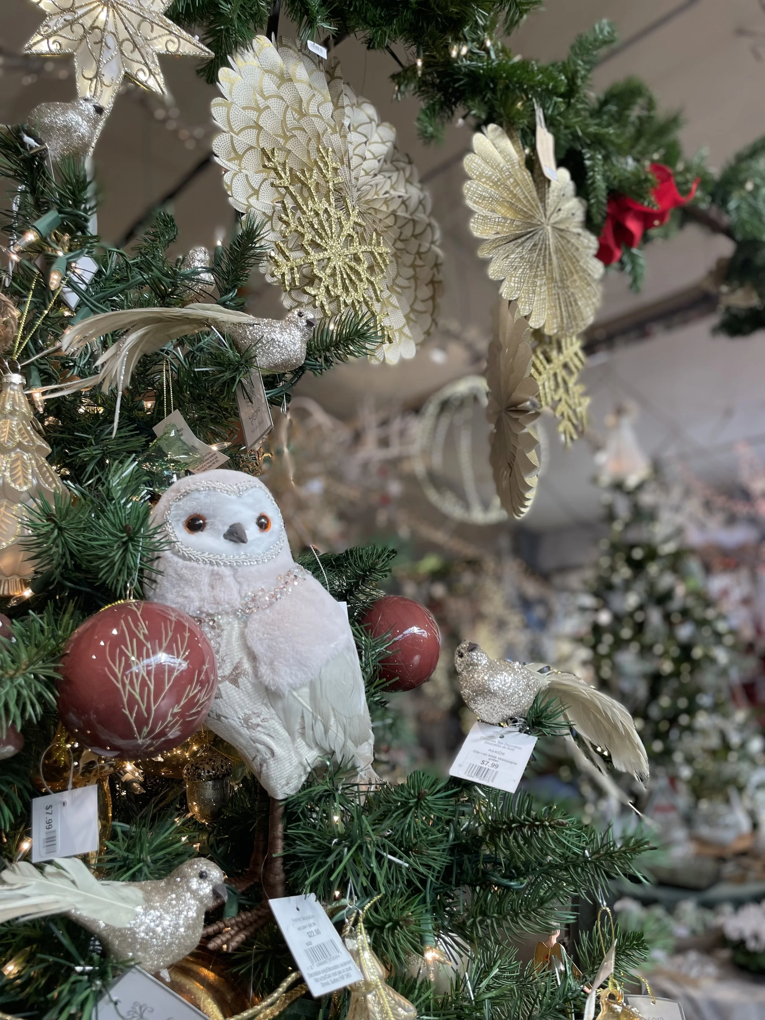 Christmas tree decorated with gold, white, and red ornaments, including an owl ornament and glittery birds, with a background of more decorated trees.
