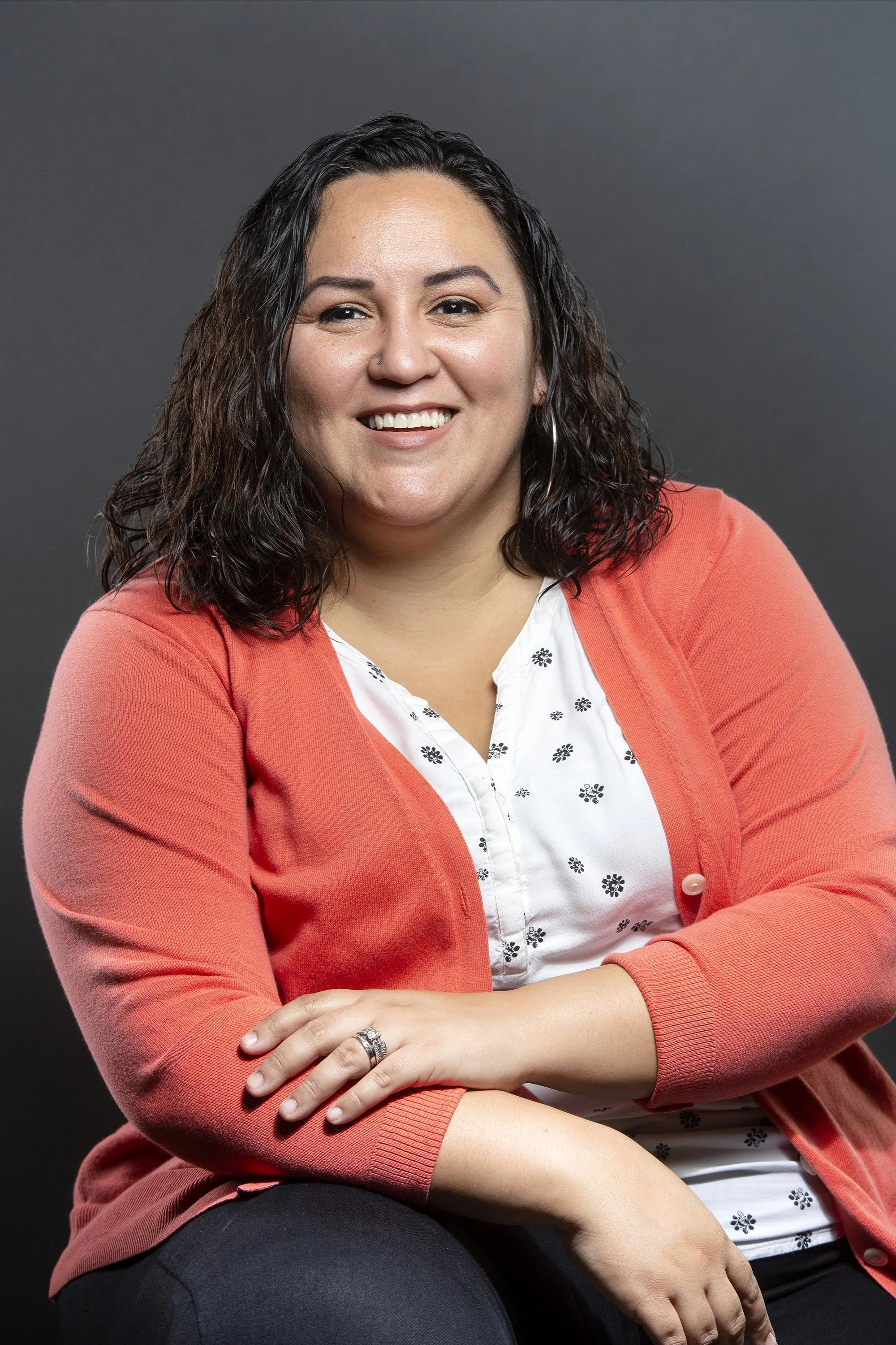 Smiling person wearing a coral cardigan and white blouse, sitting against a dark gray background.