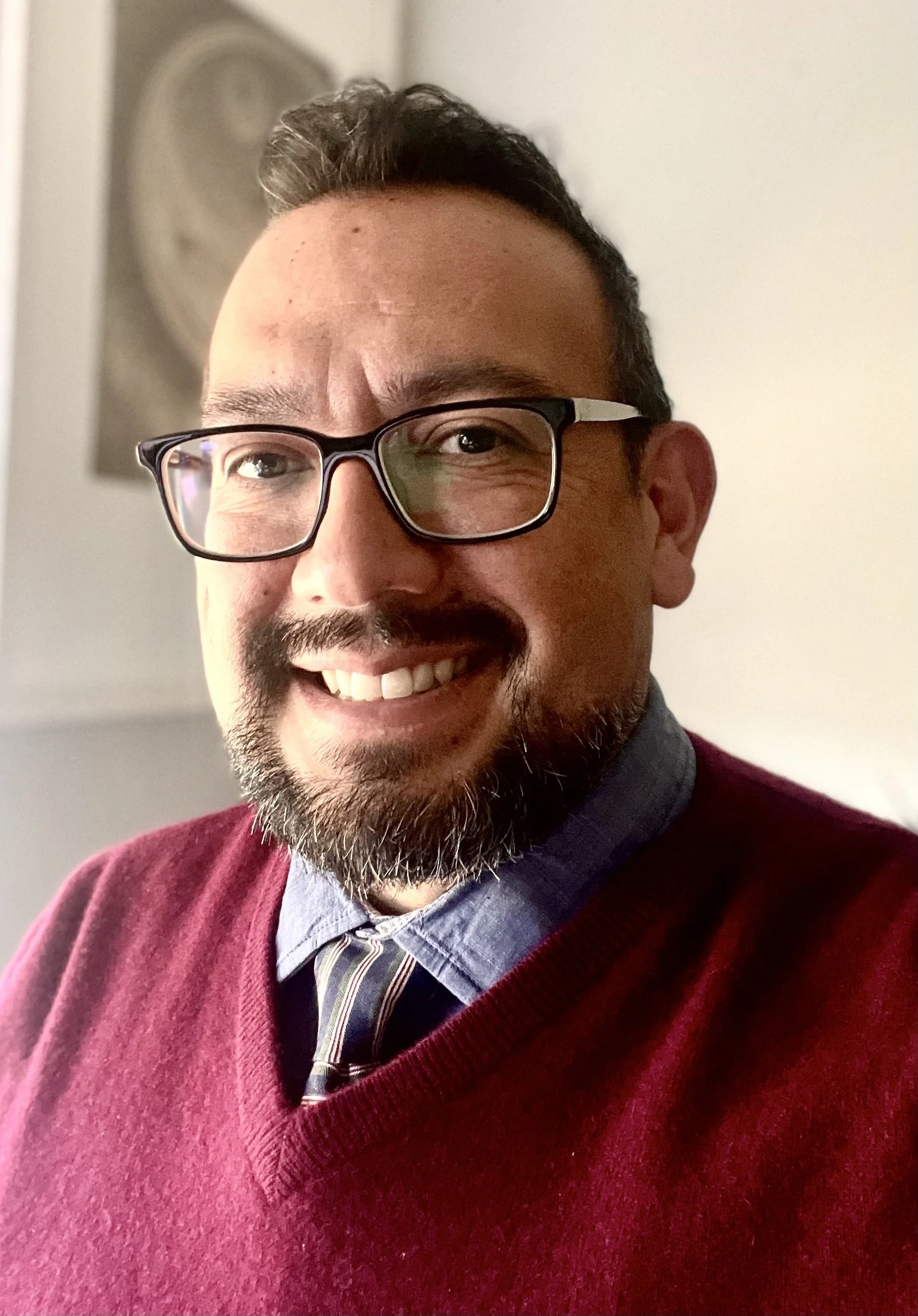 A smiling person wearing glasses, a maroon sweater, a blue collared shirt, and a striped tie, standing indoors.