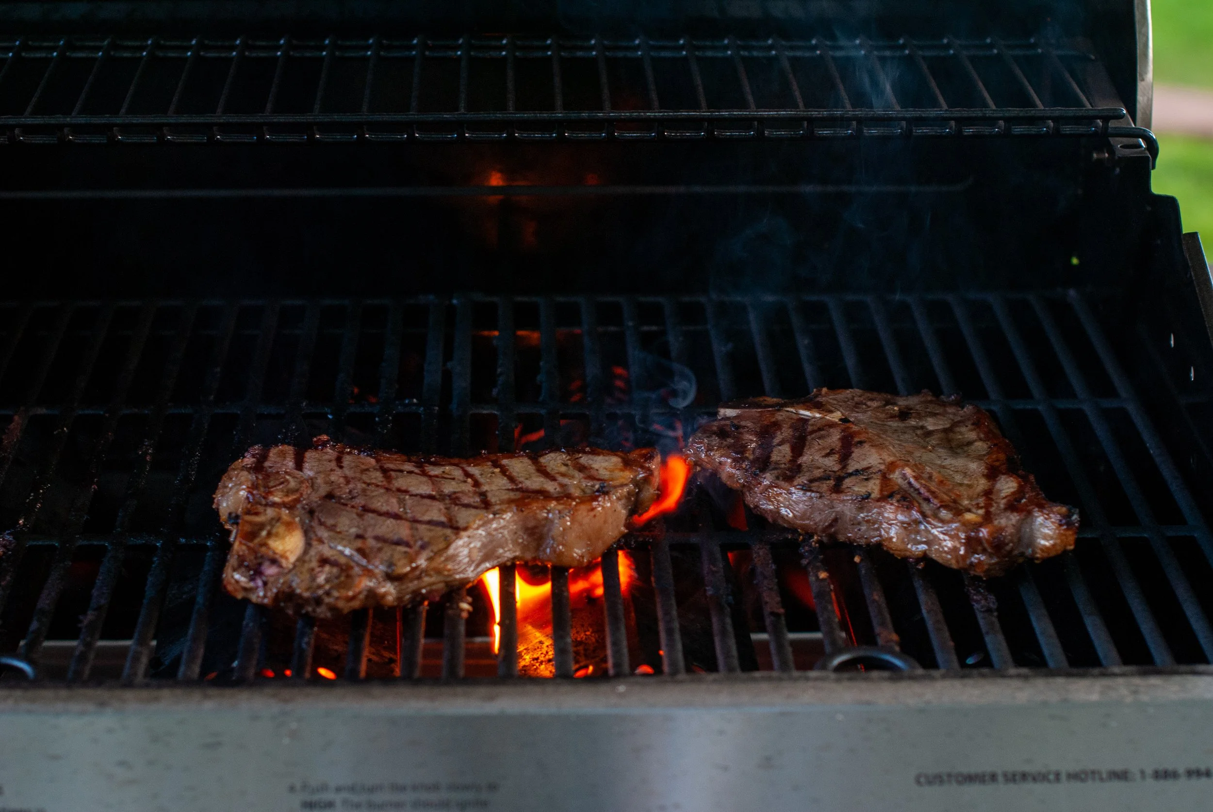 Two pieces of seasoned steak cooking on a grill over hot coals, with grill grates visible and some open flame beneath.