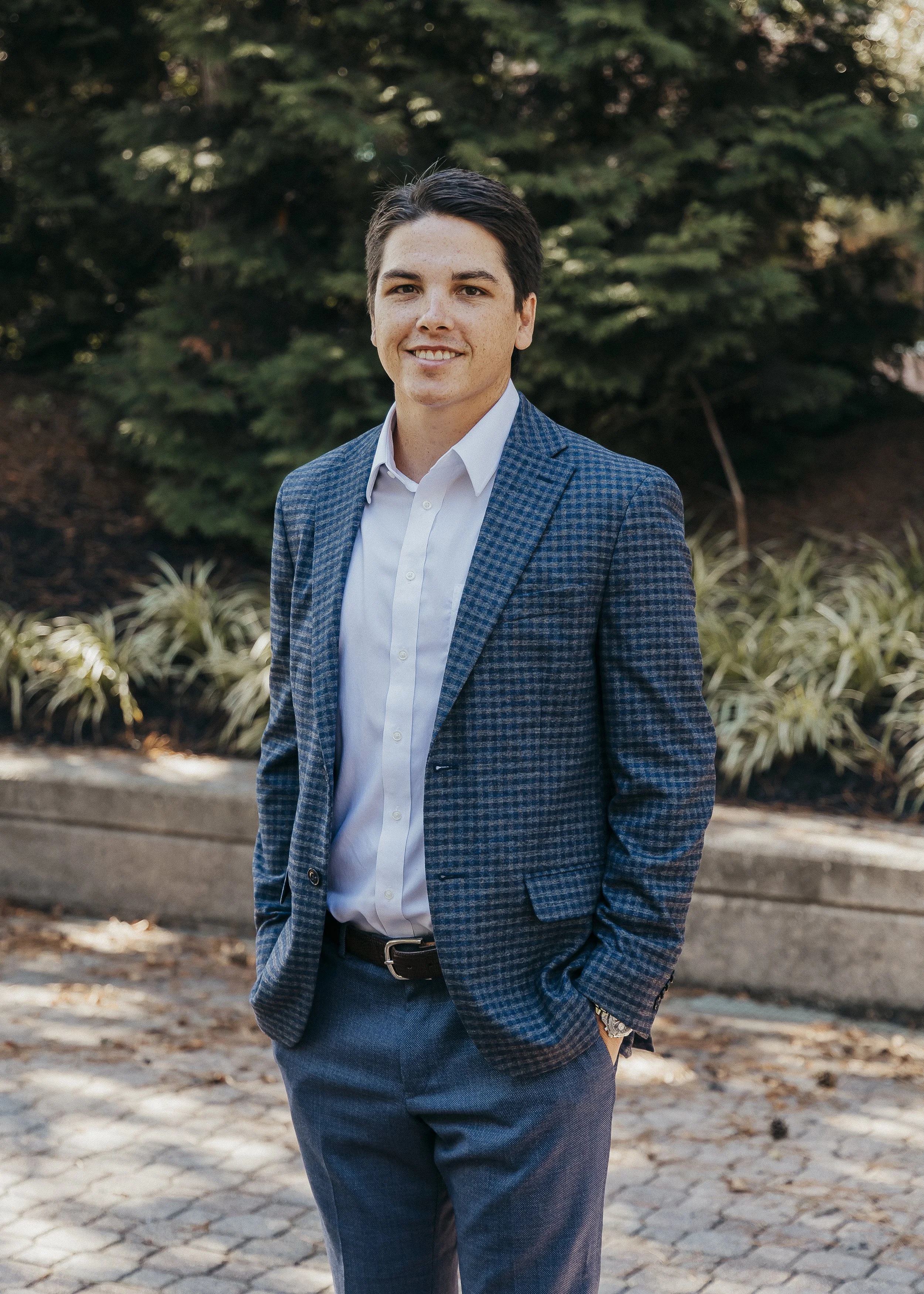 A man with dark hair wearing a white shirt and blue plaid blazer standing outdoors in front of green bushes and trees, smiling with one hand in his pocket.