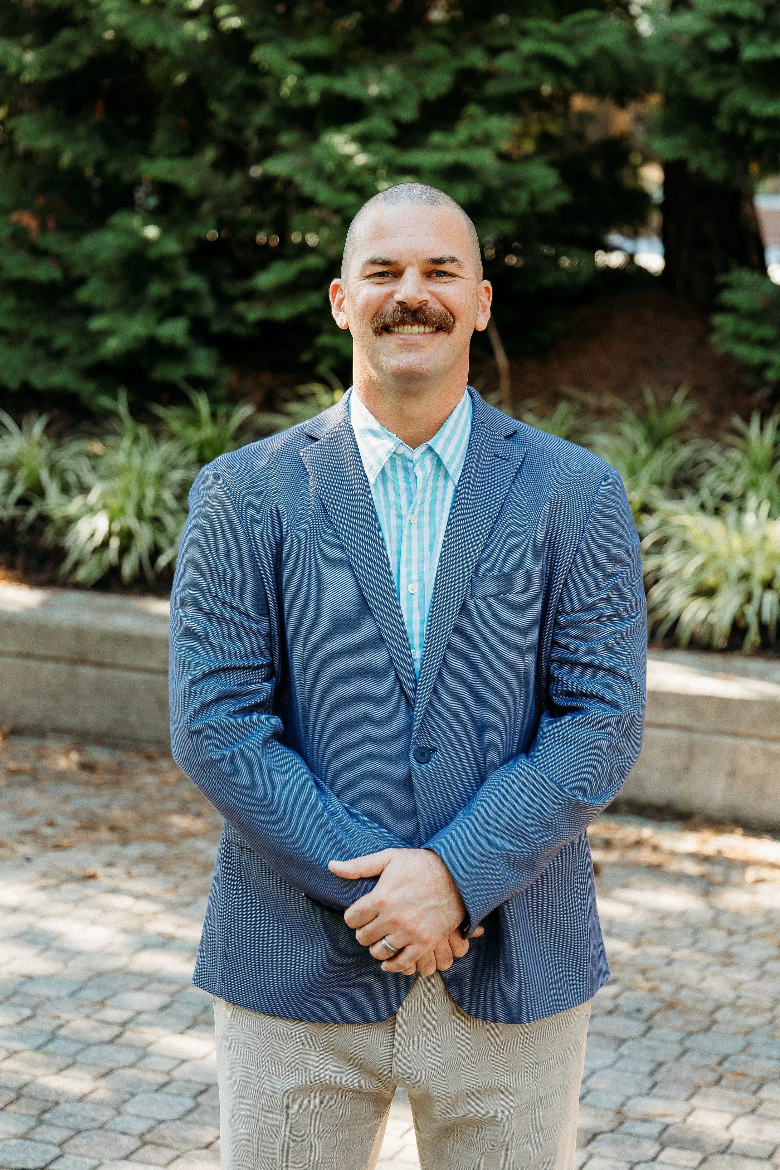 A man with a bald head and mustache, wearing a blue plaid blazer over a striped dress shirt, standing outdoors with trees and plants in the background.