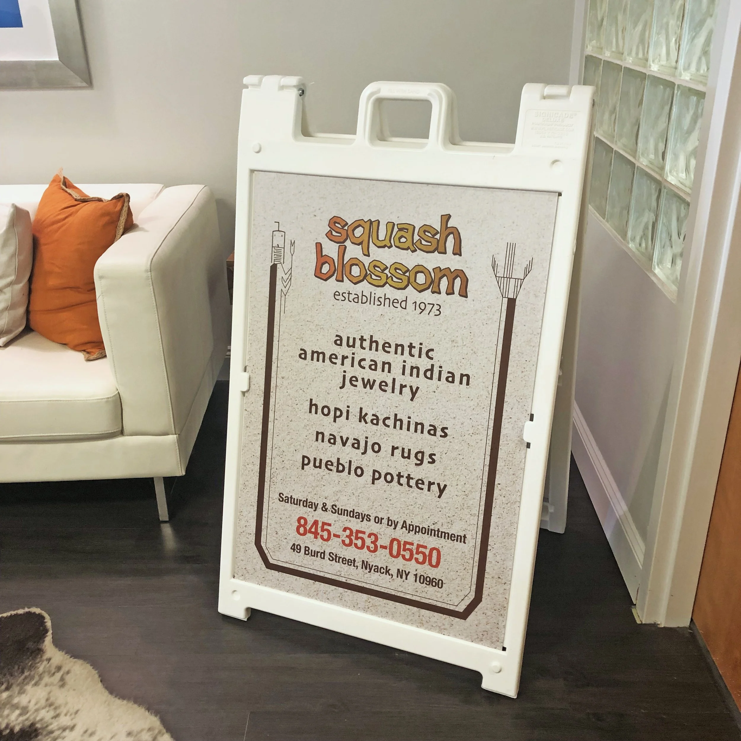 Signboard for Squash Blossom, established 1973, with details about authentic Native American jewelry, pottery, rugs, and pueblo items, on a sidewalk at 49 Burd Street, Nyack, NY.