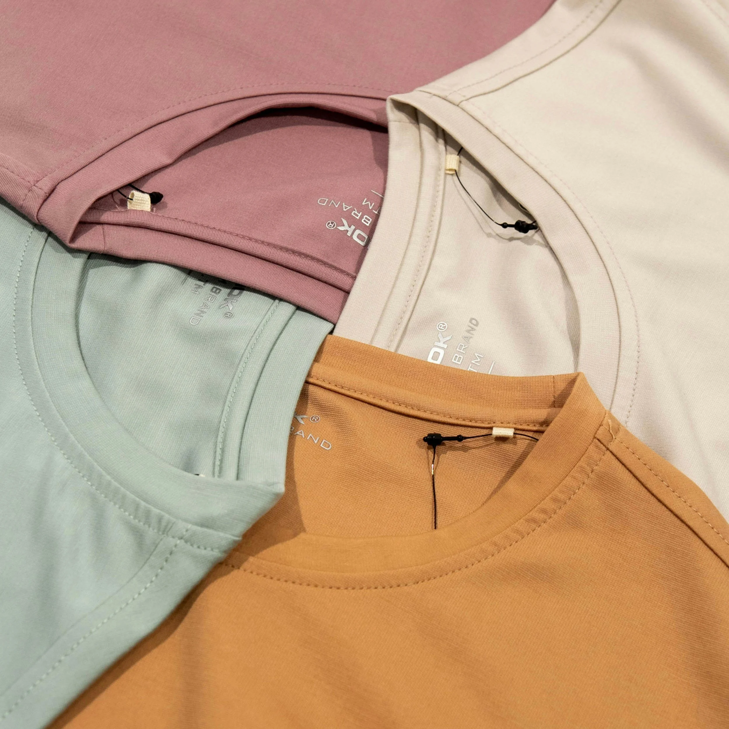 Close-up of folded pastel-colored T-shirts in pink, cream, mint green, and beige, with visible tags and stitching.