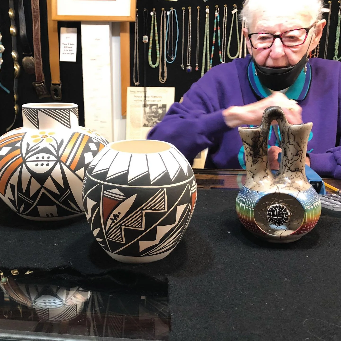 Three decorated pottery vases with black, white, and orange patterns on a black cloth, with a person wearing glasses and a mask behind them, sitting at a table with jewelry displayed on the wall behind.