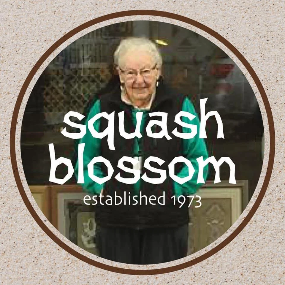 A woman with glasses smiling, standing in front of a display of plants at Squash Blossom, established in 1973.