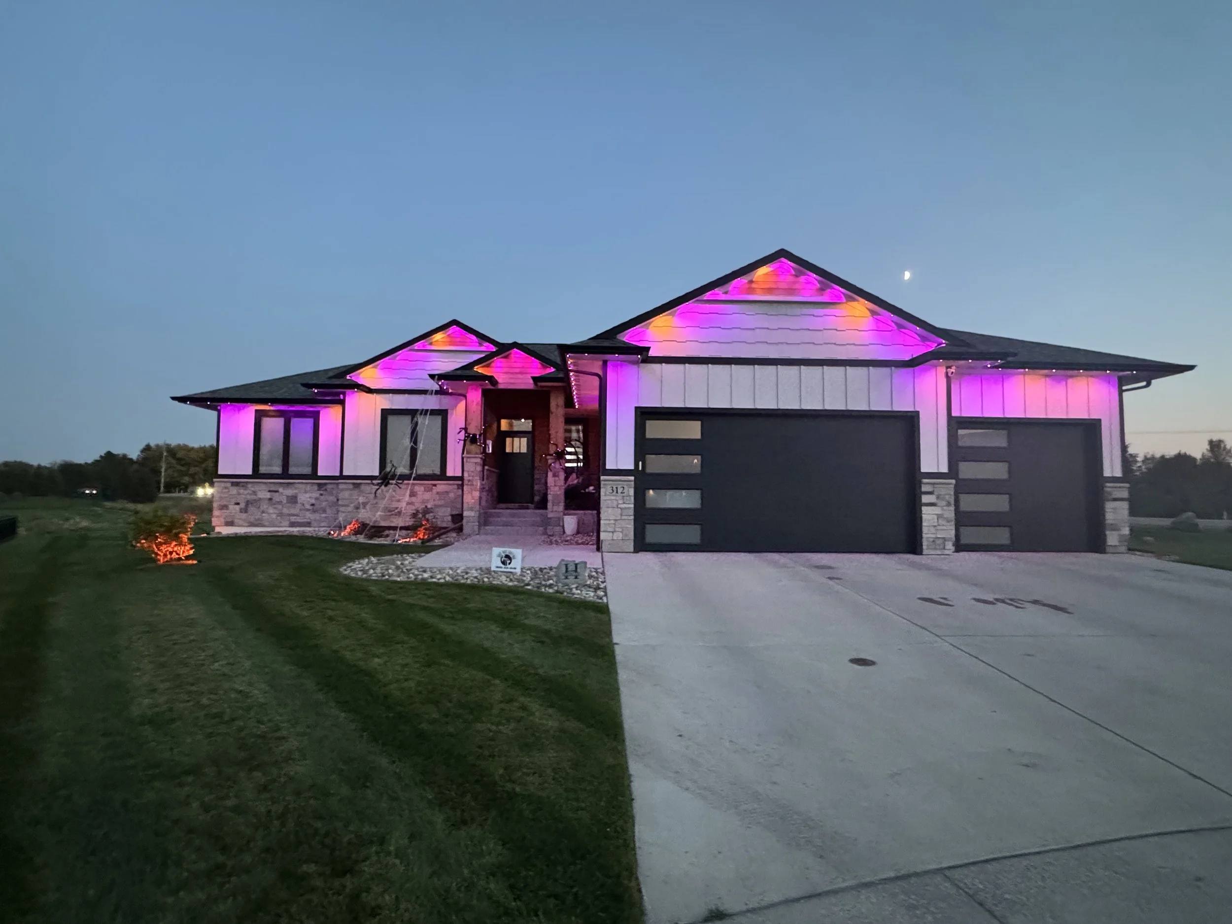 Modern house with exterior lights featuring pink and purple hues, large driveway, and landscaped lawn at dusk.