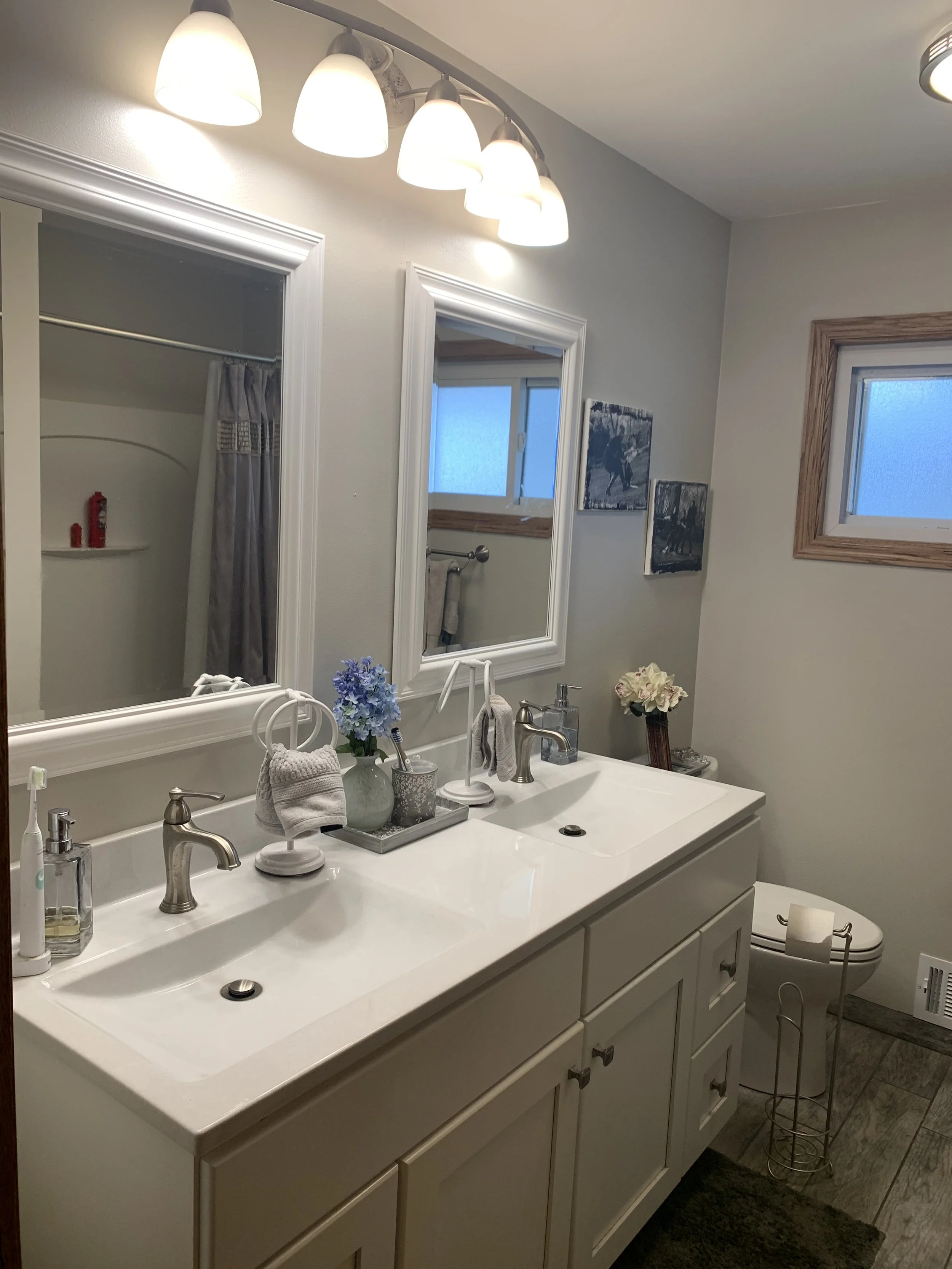 Modern bathroom with double sink vanity, mirrors, and overhead lighting. Decor includes flowers and towels. There's a window and a glimpse of a shower curtain.