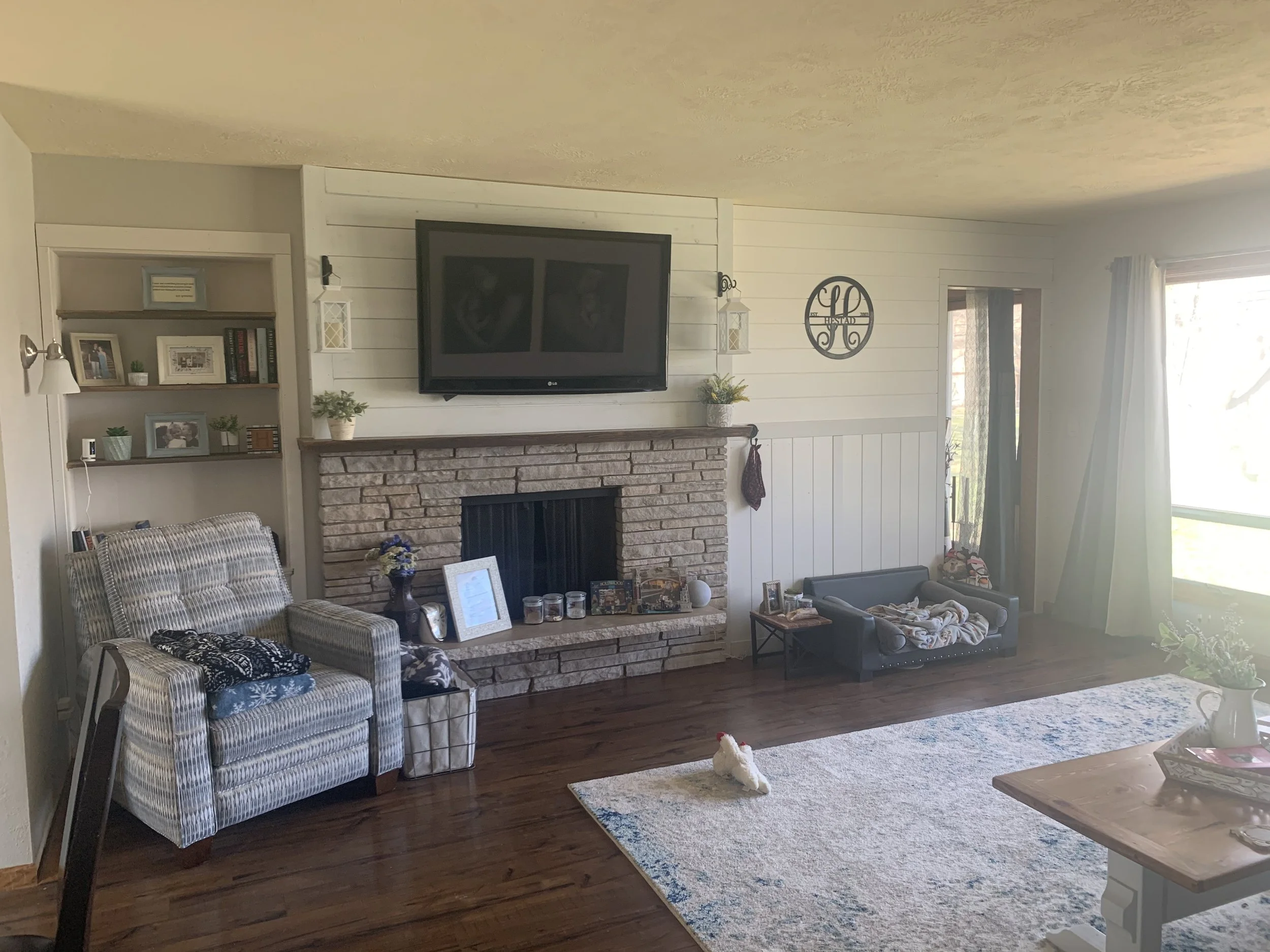 A cozy living room with a brick fireplace and a TV mounted above it. There is an armchair with a patterned cover next to a basket and a small bookshelf with family photos and decorative items. The room has a large rug on polished wooden floors, and a small dog bed with a blanket in the corner. A window with curtains lets in natural light.