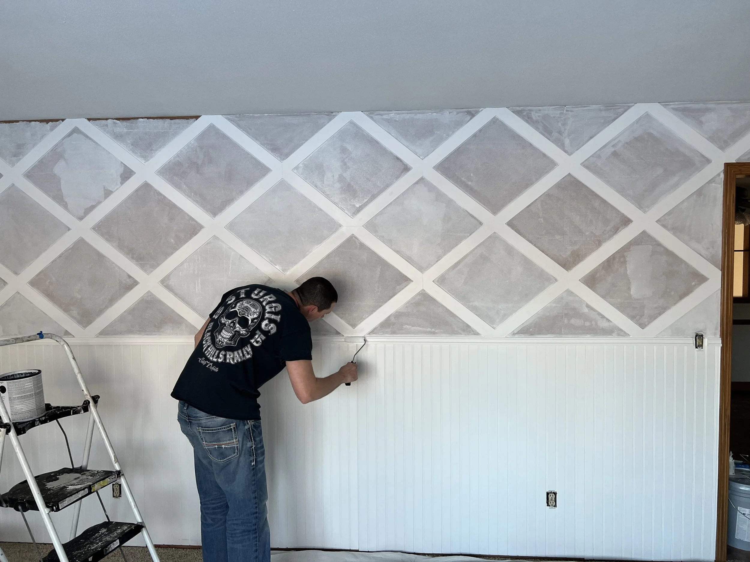 Person painting a wall with a roller in a room, featuring a diamond pattern on the upper half and white wainscoting on the lower half. A step ladder and paint can are nearby.