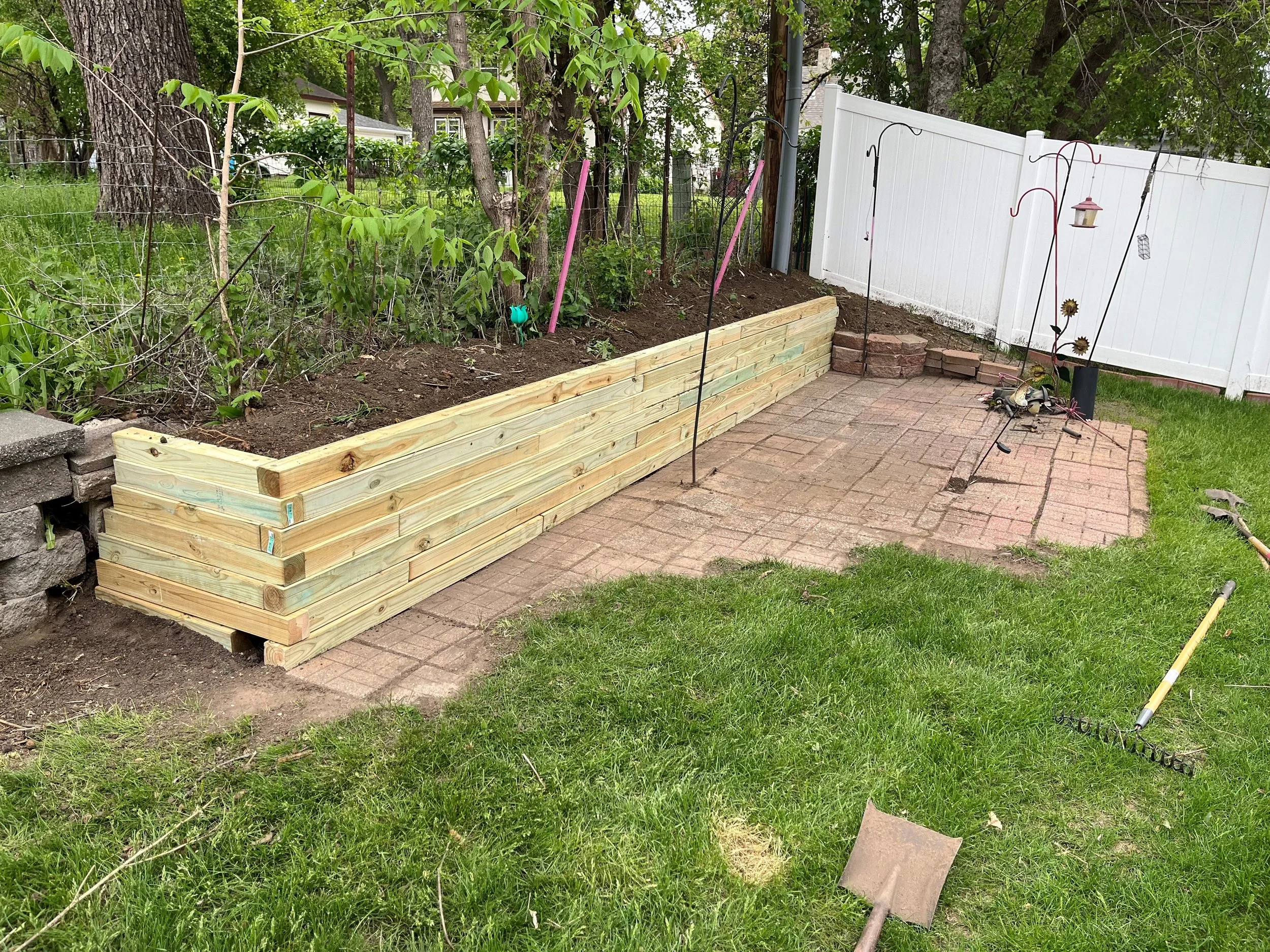 Garden area with a raised wooden planter box, surrounded by a patio and white fence, with gardening tools and plants visible.