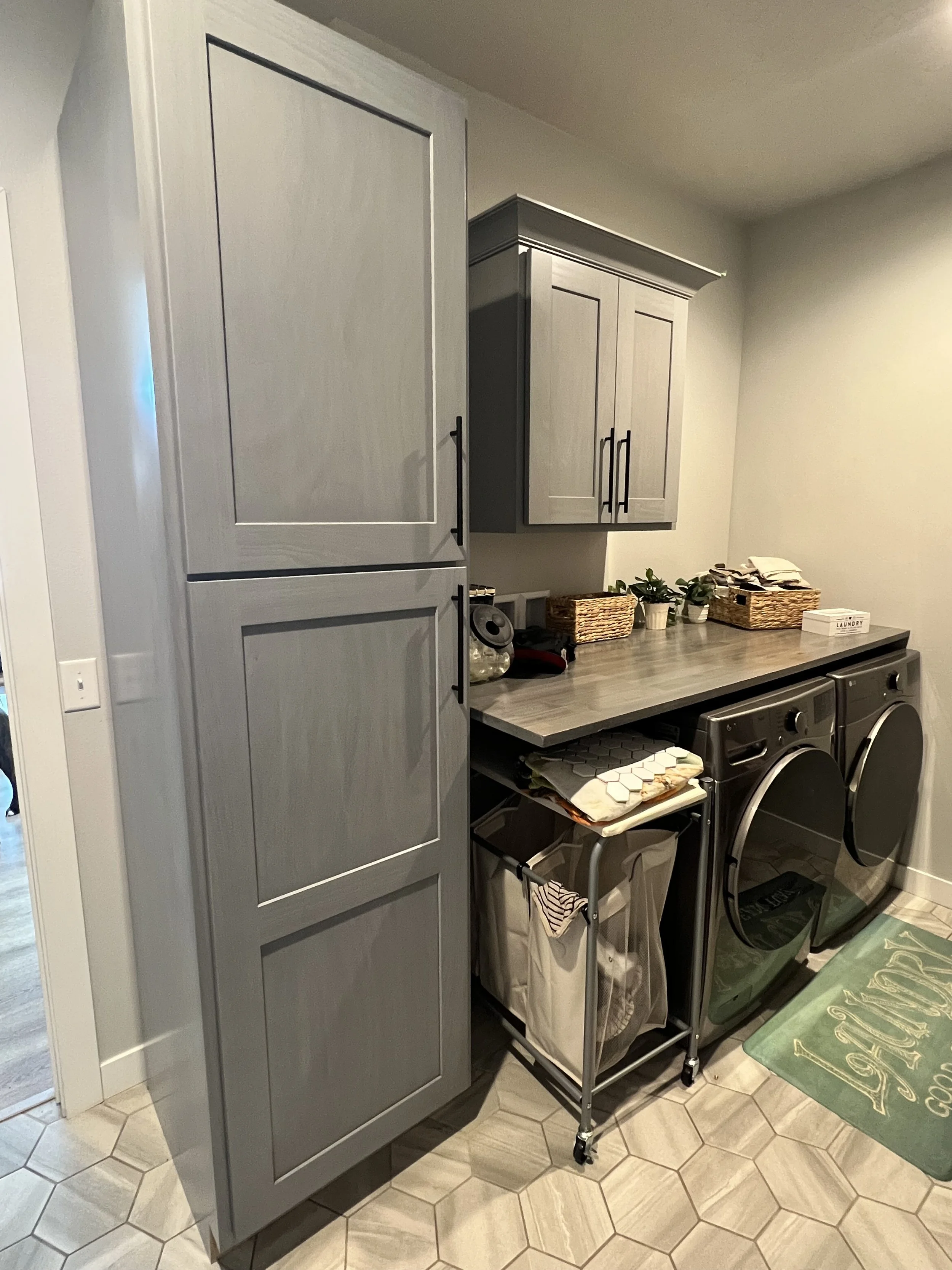 Laundry room with gray cabinets, a washer and dryer, and a folding counter. Laundry baskets on wheels and storage baskets are used for organization. A "Laundry" rug is on the floor.