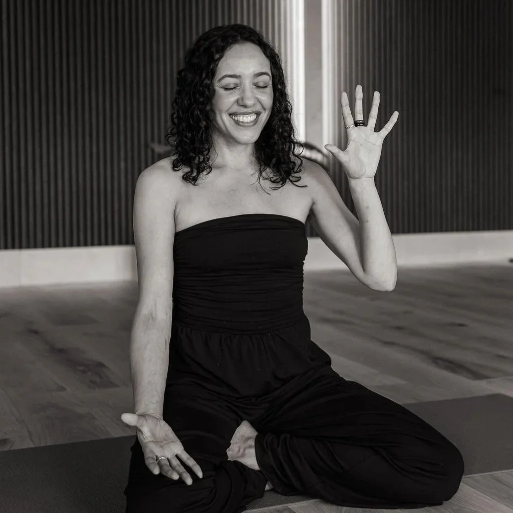 Black-and-white photo of a woman in workout clothes sitting on the floor, stretching her leg, smiling, with curly hair and a playful expression.