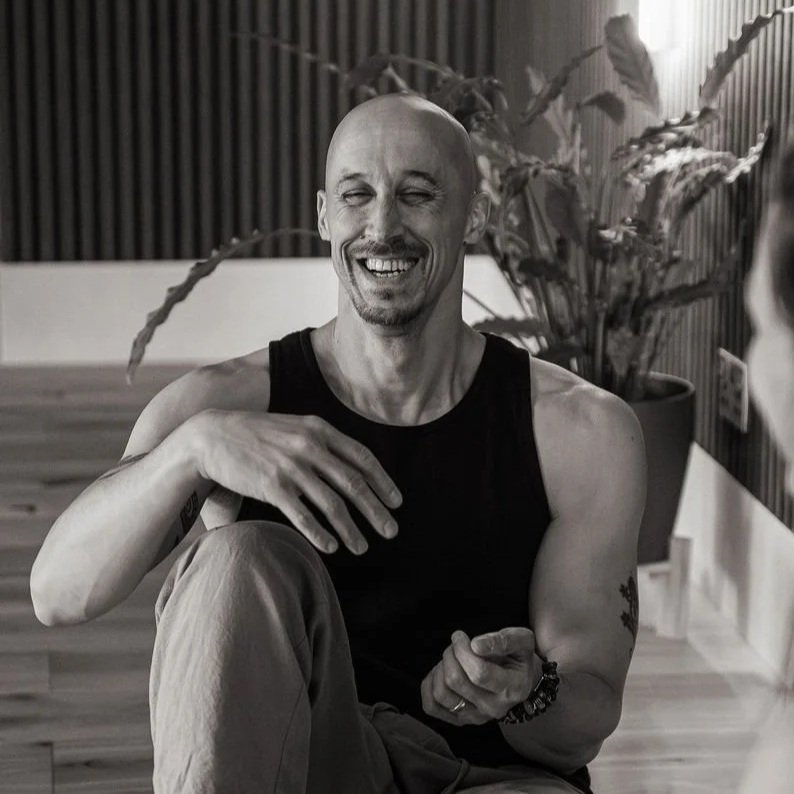 Black and white photo of a bald man smiling, looking to the side, wearing a sleeveless top.
