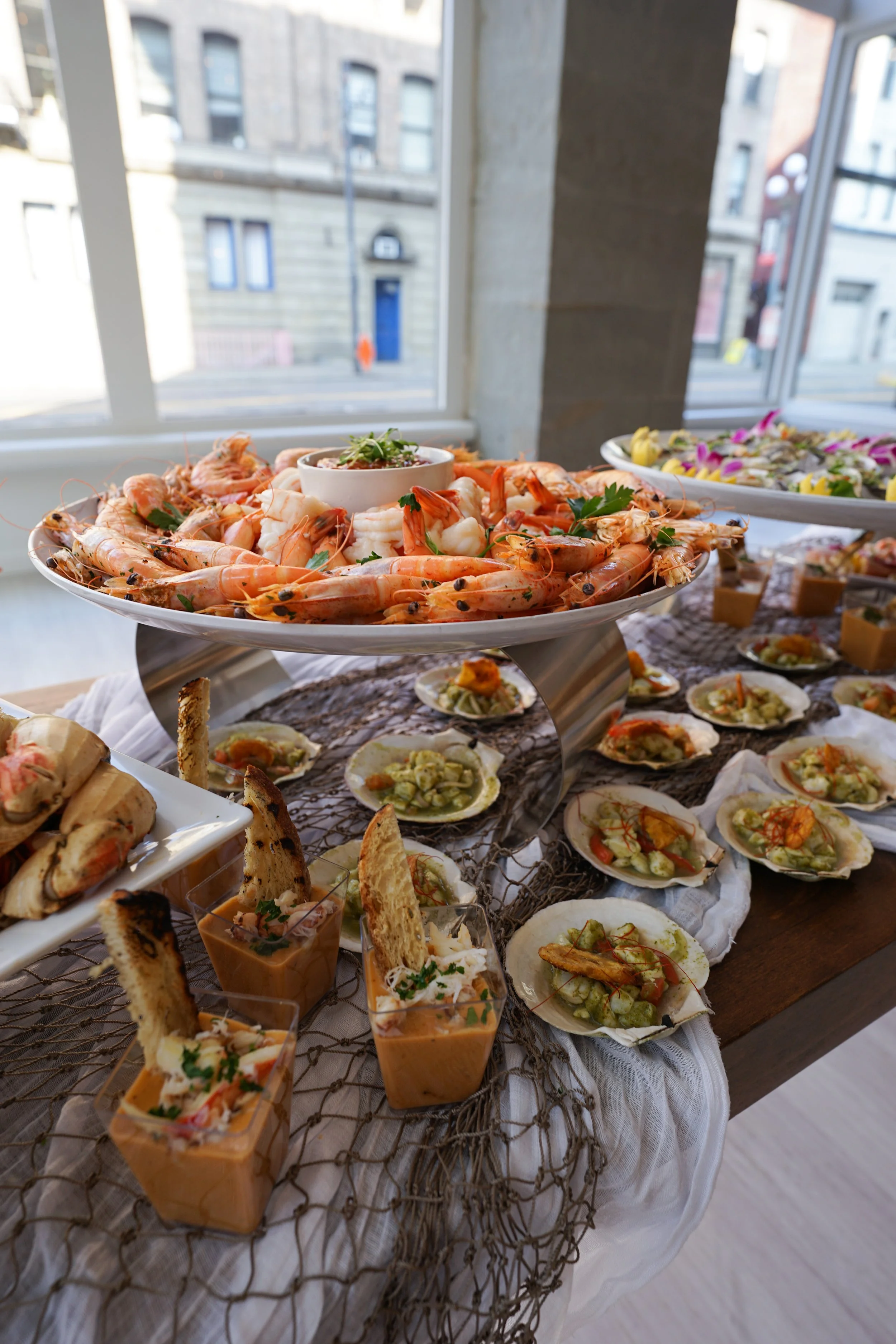 A seafood buffet with shrimp, oysters, and small appetizer cups on a table near a large window.