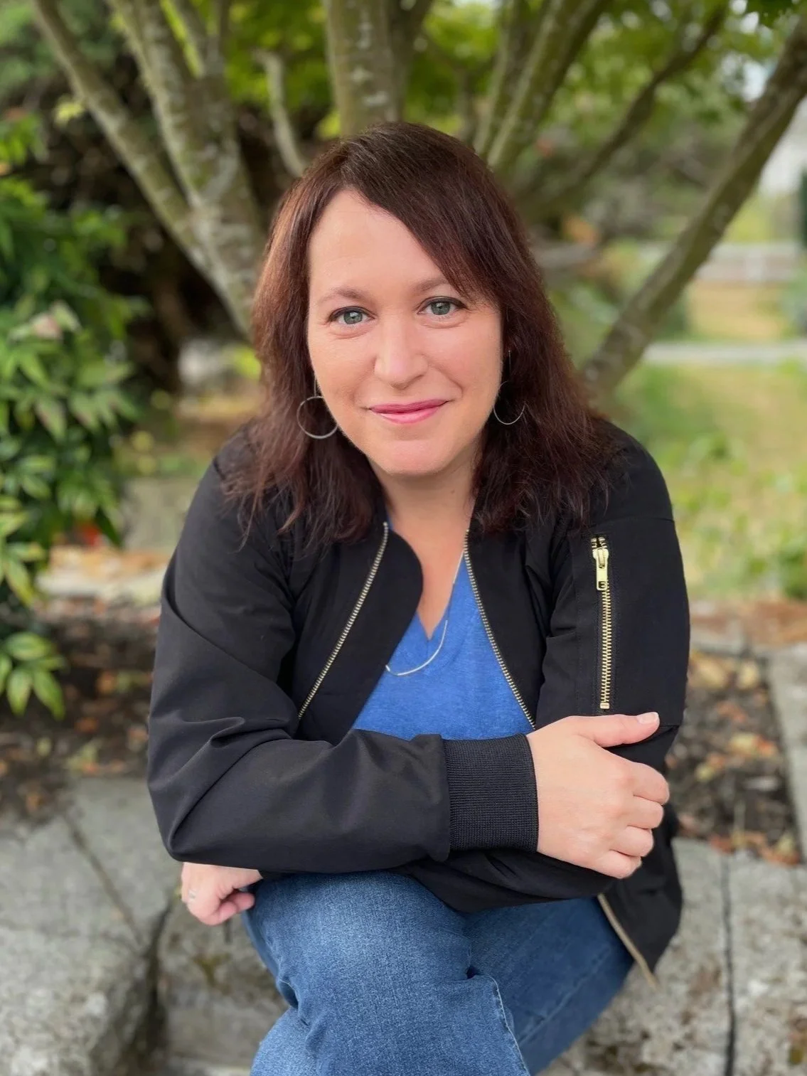 A woman with shoulder-length brown hair, smiling, wearing a black denim jacket and a teal top, standing outdoors with a blurred green background.