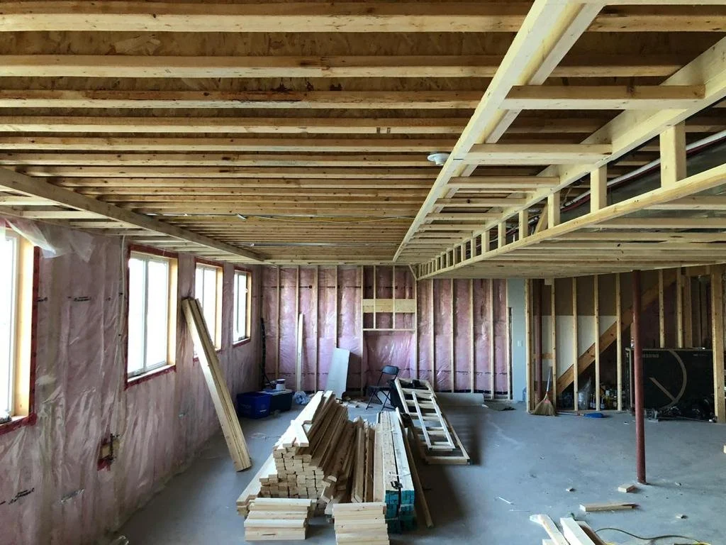 Interior of a house under construction with exposed wooden framework, pink insulation, and construction materials on the floor.