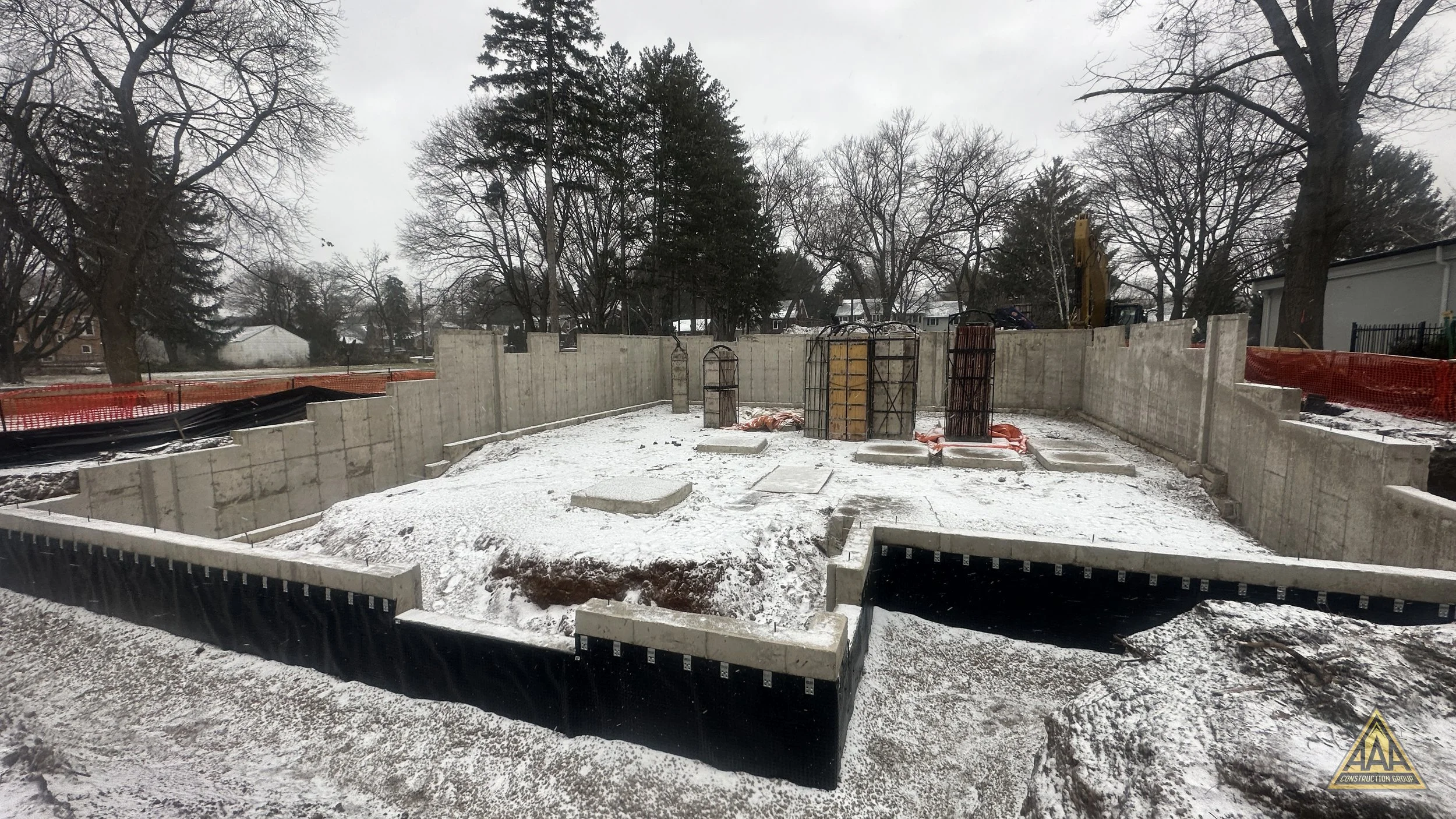 Construction site with concrete foundation walls and rebar protruding for a building, snow on the ground, surrounded by trees and overcast sky.