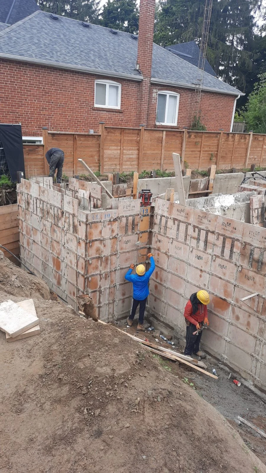 Construction workers building a brick foundation wall in a backyard, with a wooden fence and brick house in the background.