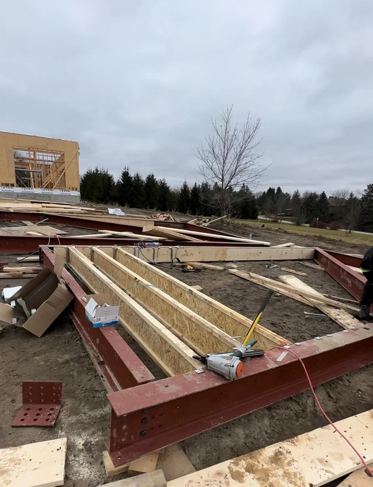 Construction site with a building frame under construction, with wooden beams and steel beams, on a cloudy day and some trees in the background.