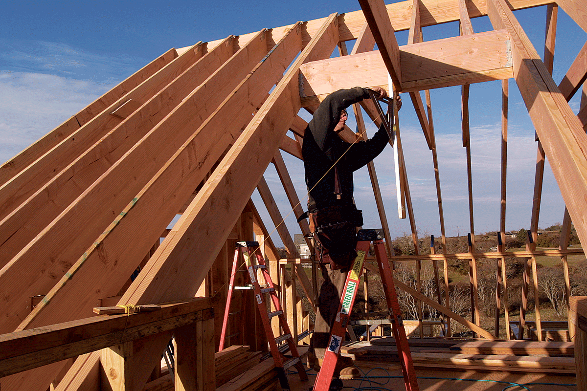 A person working on framing the roof of a house under construction, standing on a ladder and using a measuring tool.