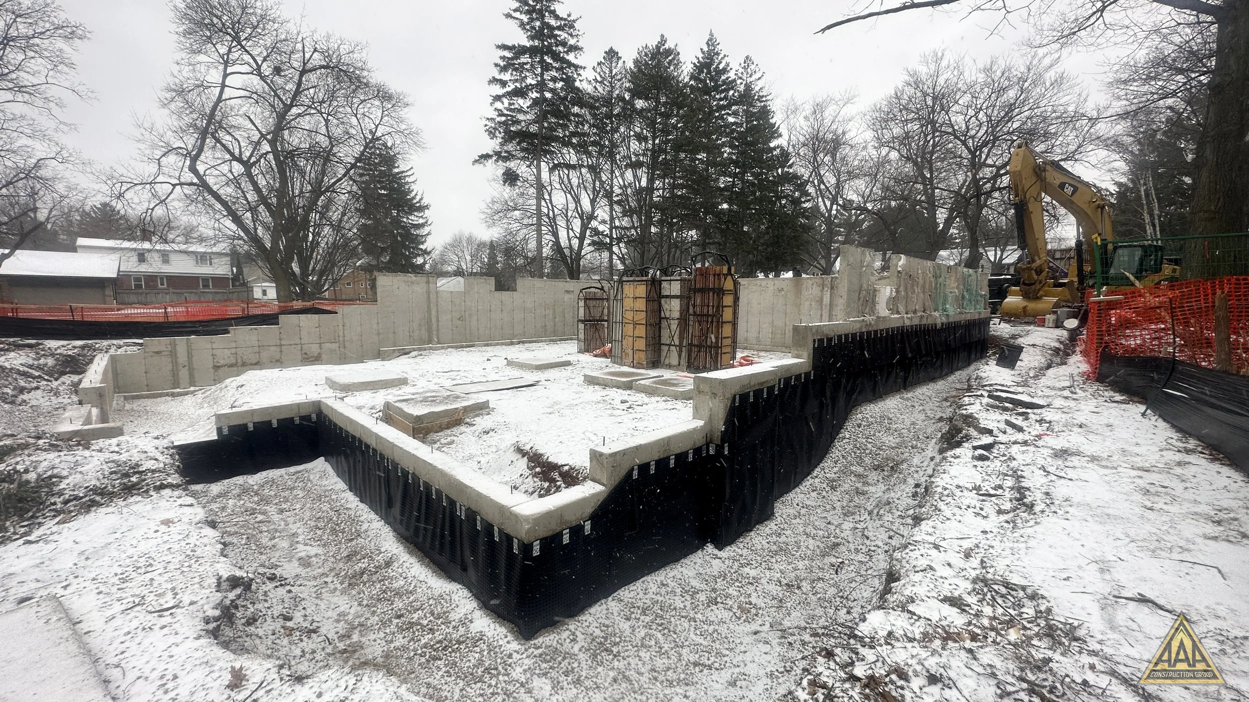 Construction site with concrete foundation and exposed rebar, surrounded by snow and trees, with a yellow excavator on the right side.