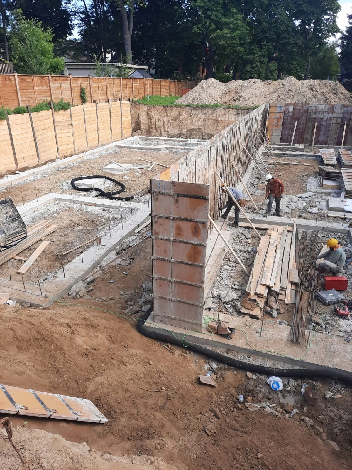 Construction site with workers building a foundation, wooden forms, steel rebar, and dirt piles in the background.