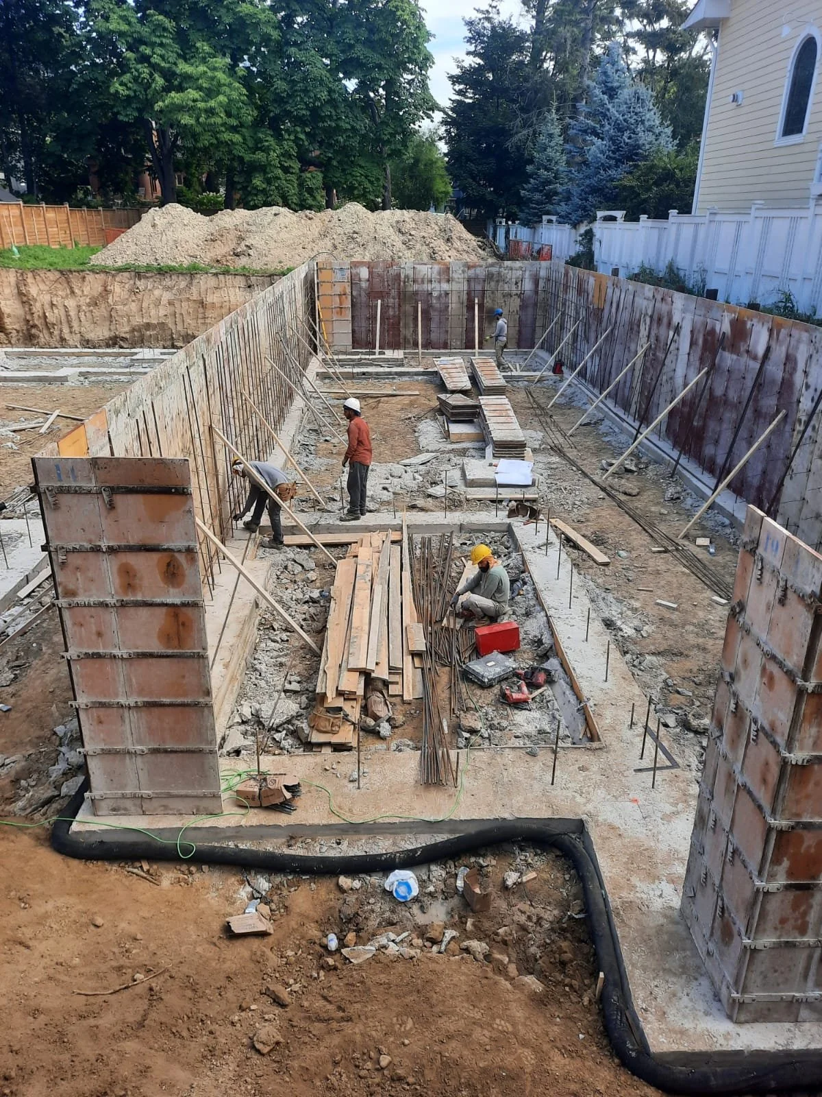 Construction workers building foundation with steel reinforcement and wooden formwork on a construction site, surrounded by earth and trees.