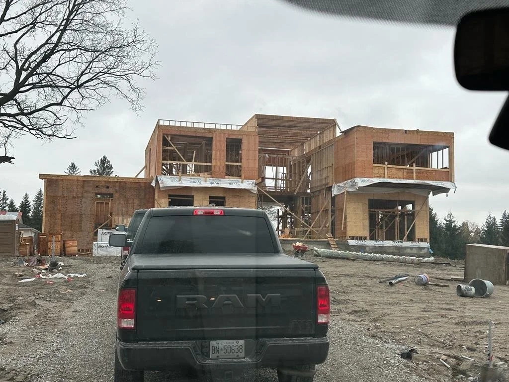 A house under construction with wooden framing, viewed from behind a black RAM truck on a gravel lot, with scattered construction debris and an overcast sky.