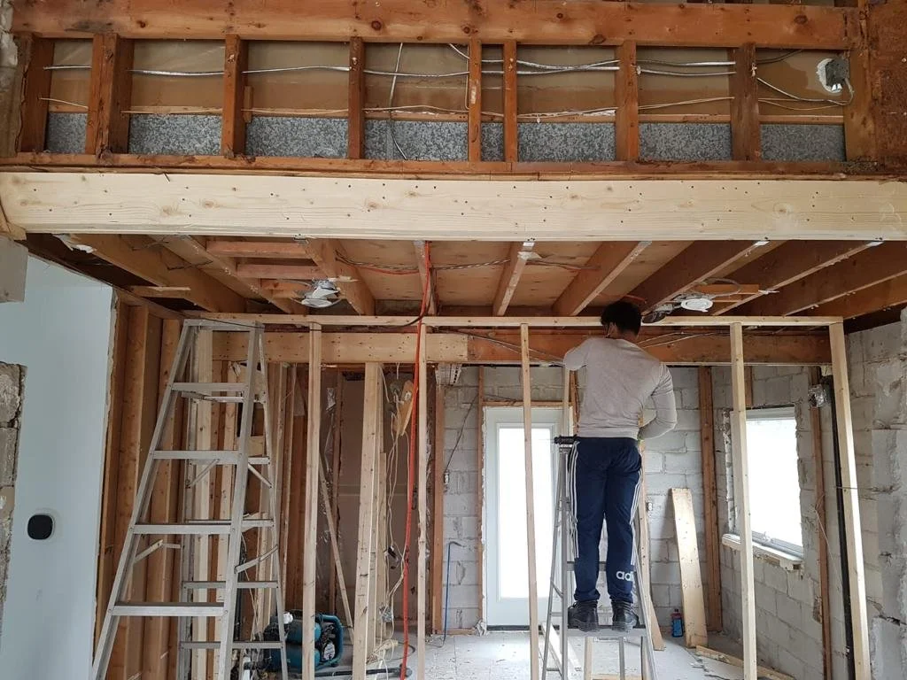 A person standing on a ladder inside a room under construction, working on the ceiling framing with exposed wooden beams and electrical wiring, with unfinished walls and a window in the background.