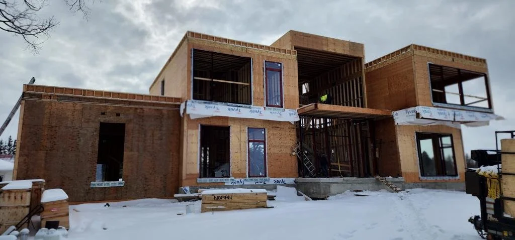 Under-construction modern house with multiple levels and large windows, partially built with wood framing, in snowy weather.