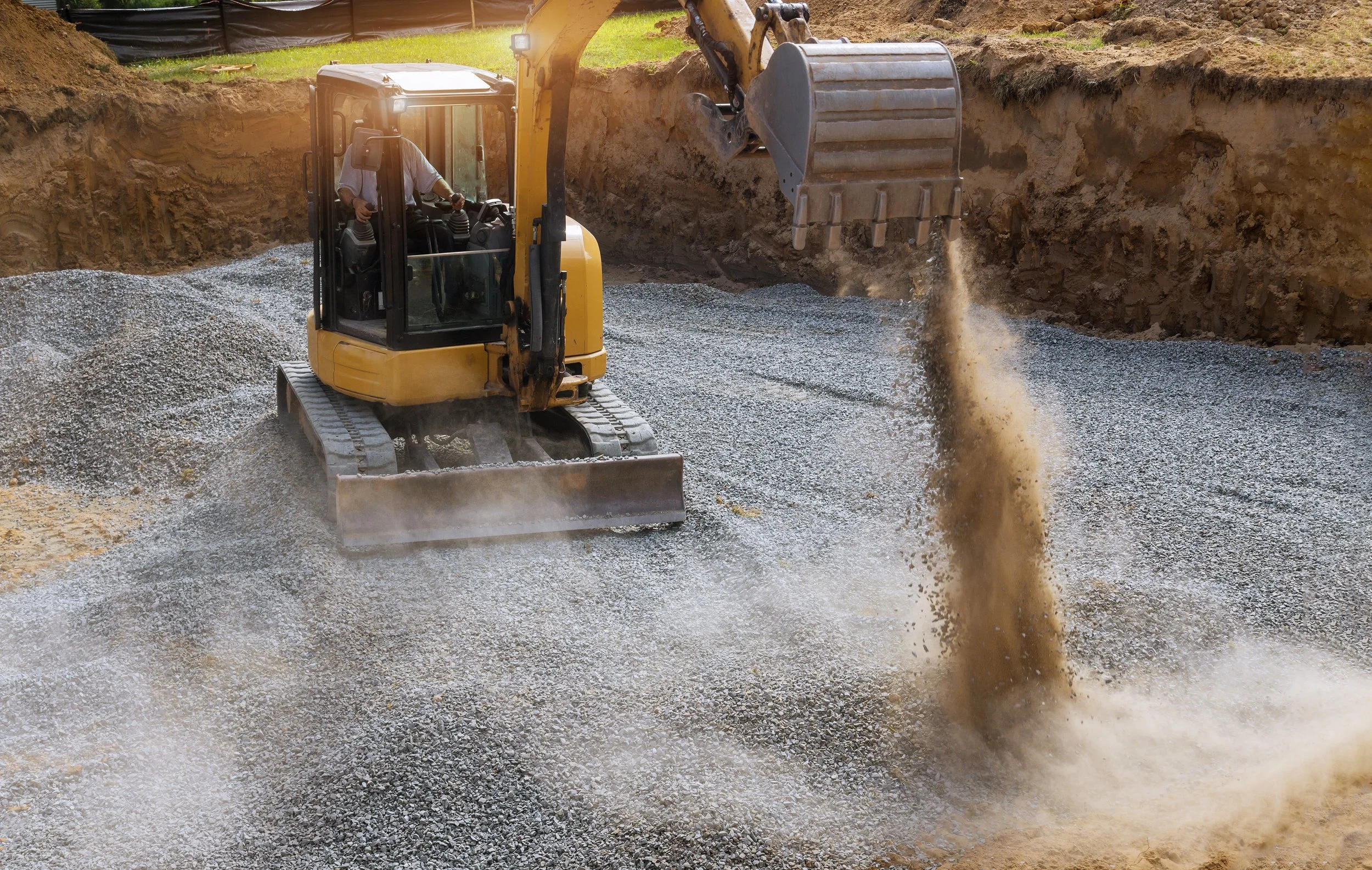 A yellow excavator with a hydraulic arm and bucket is operating on a construction site, moving gravel and dirt. The excavator is on tracks and there are raised dirt walls in the background.